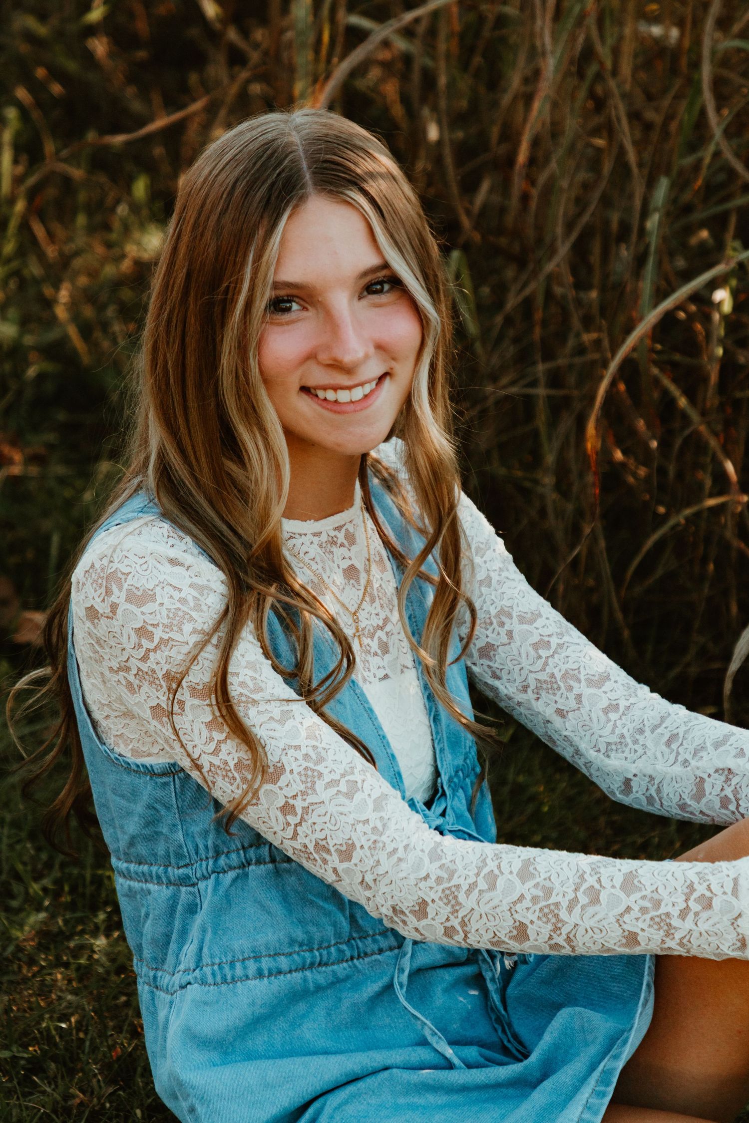 A person in a denim dress and white lace top poses outdoors against a natural autumn background.