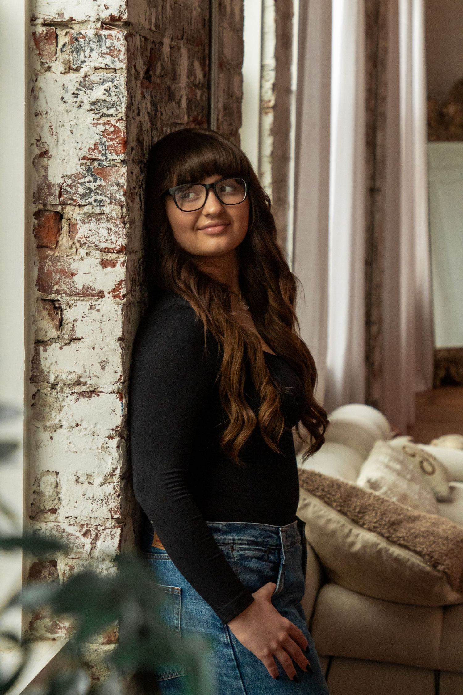 A person wearing glasses and a black long sleeve top stands by a rustic brick wall near a beige couch.