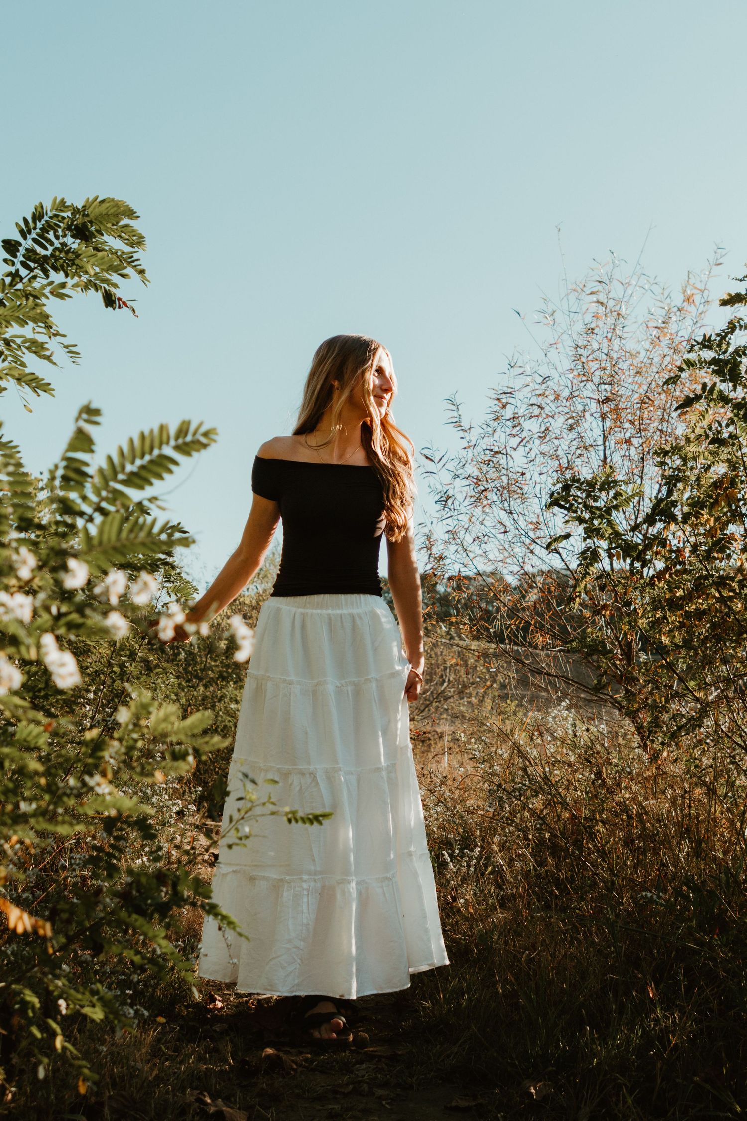 A person in a black top and white maxi skirt stands on a nature path during sunset.