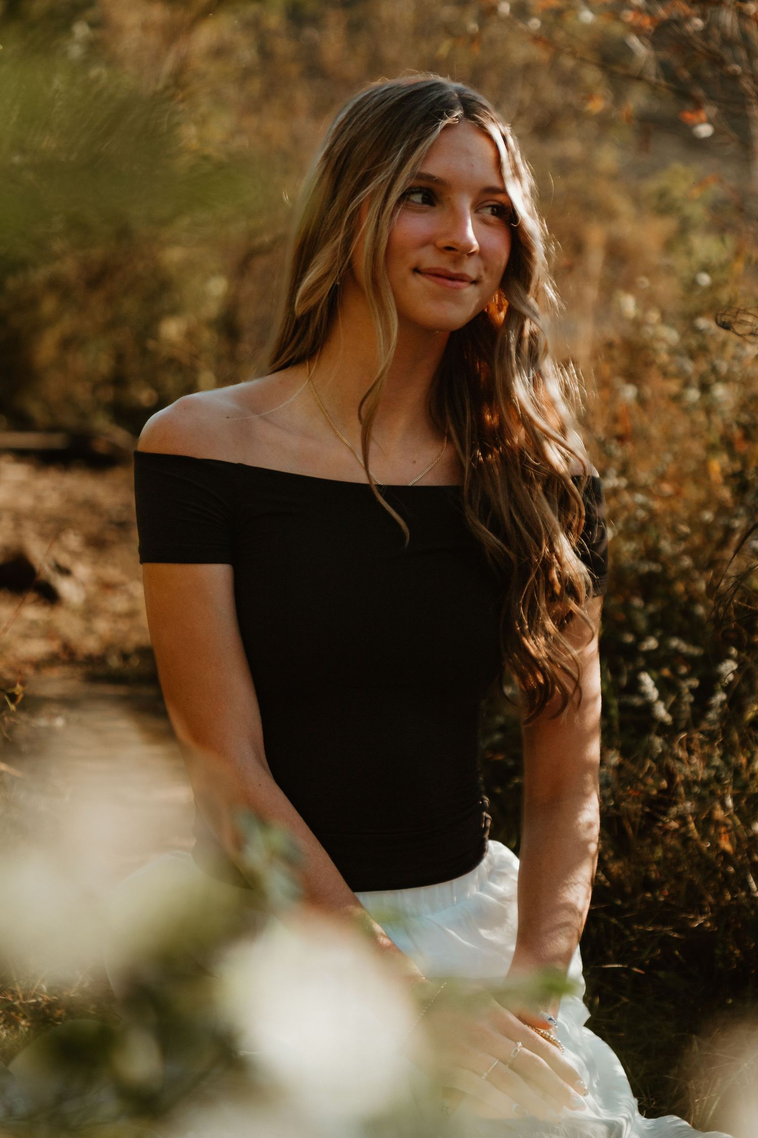 A woman in a black off-shoulder dress poses outdoors during golden hour with warm autumn lighting.