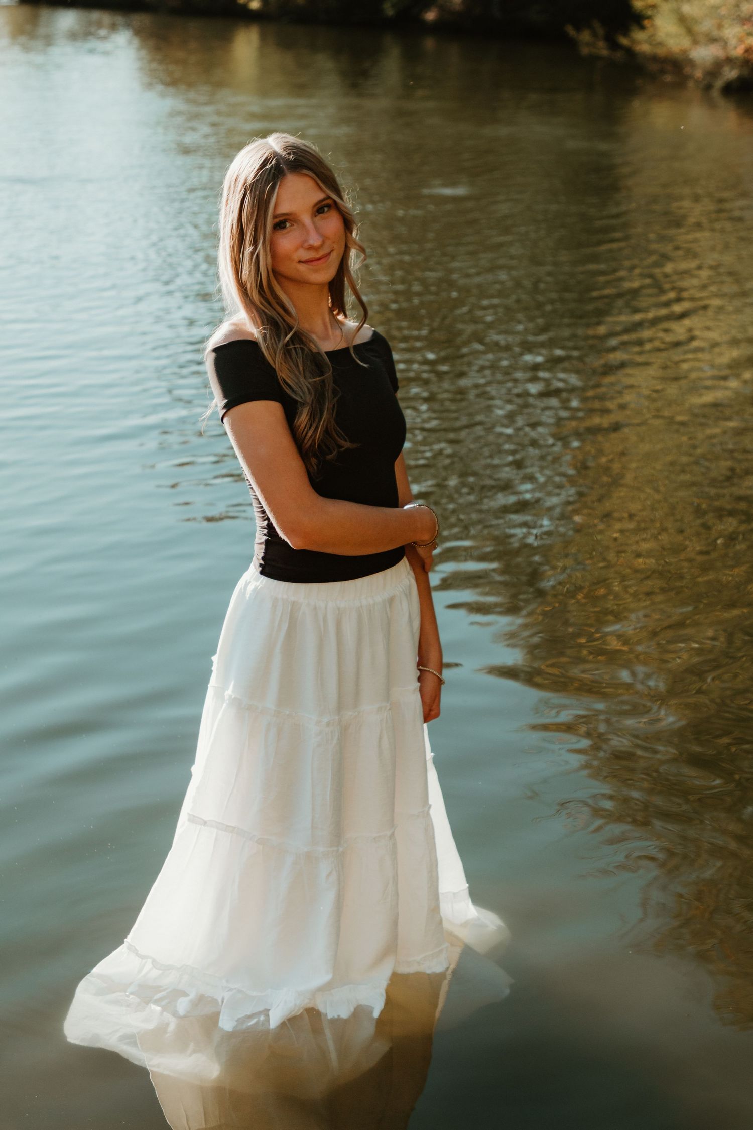 A flowing white dress contrasts with a black top as someone poses gracefully by a tranquil lake at sunset.
