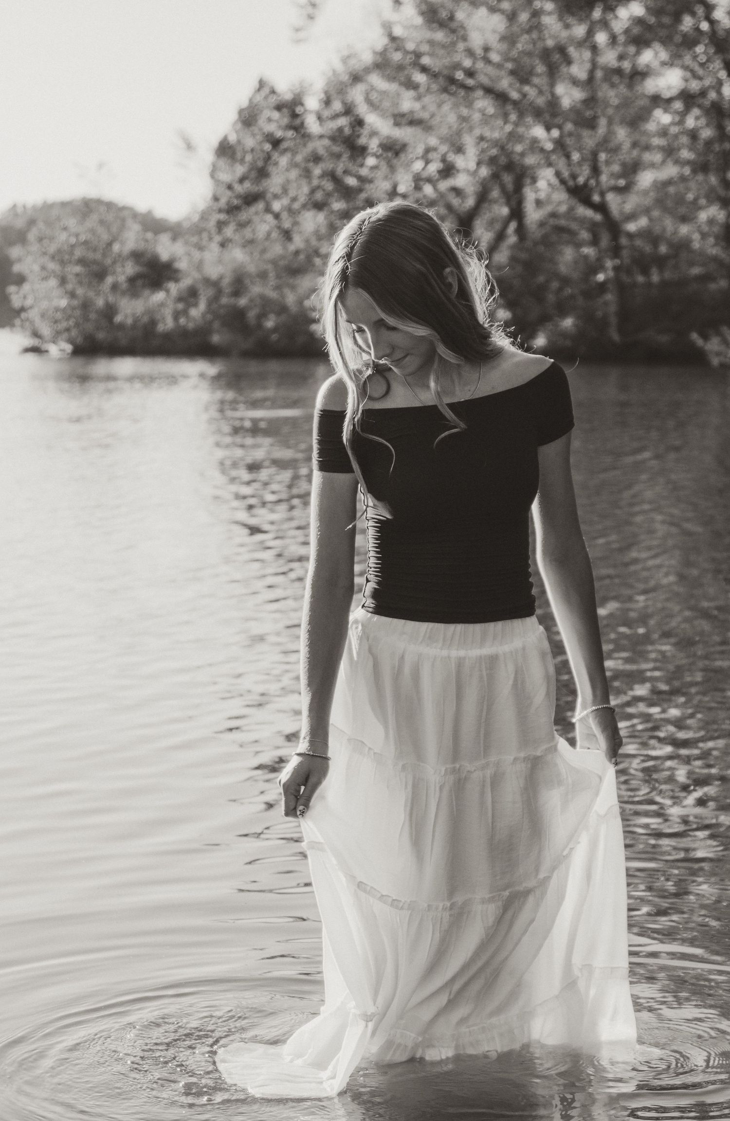 A woman in a black and white dress wades through calm water near a rocky shoreline in a dreamy monochrome scene.