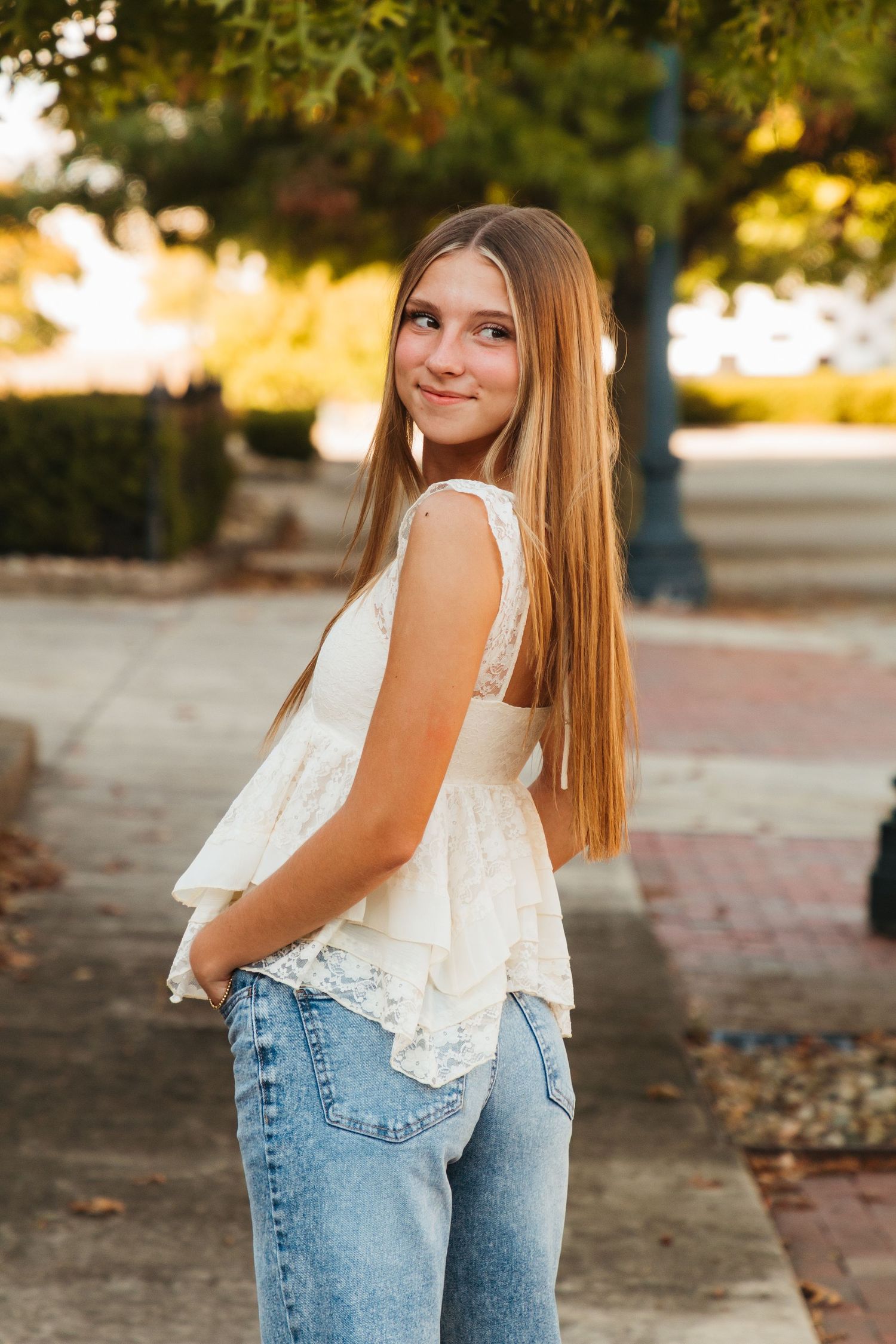 A person in a white lace top and blue jeans poses outdoors during golden hour lighting.
