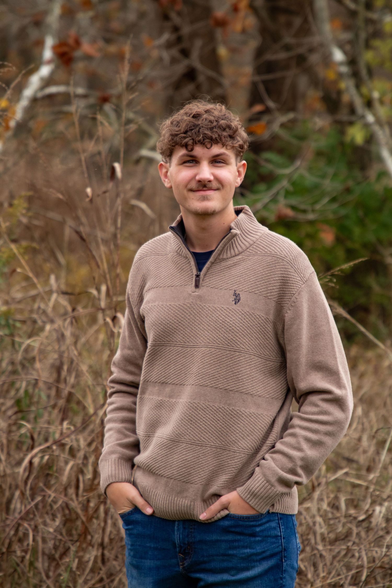 A person in a beige quarter-zip sweater and blue jeans poses outdoors against a natural autumn background.