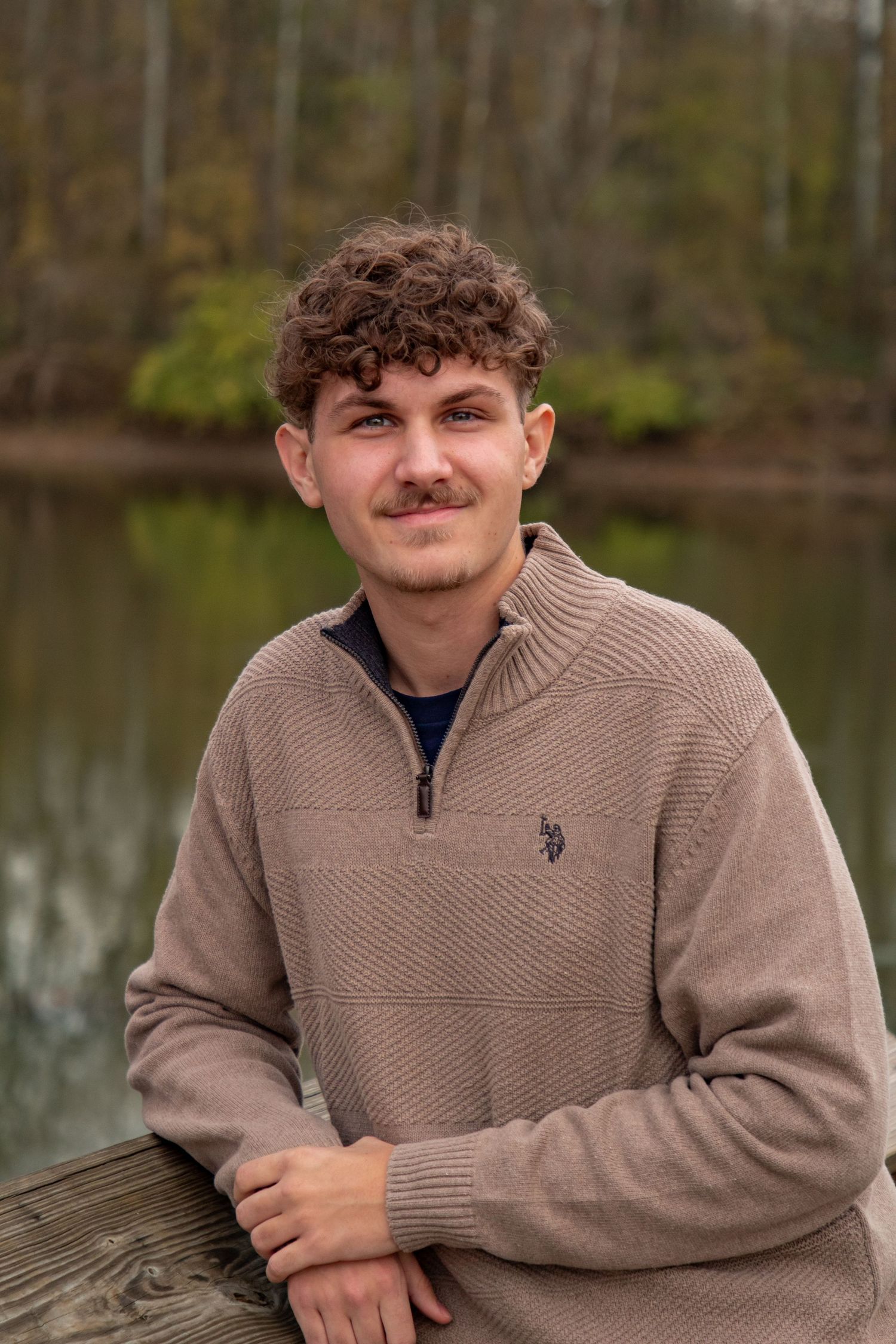 A person in a beige quarter-zip sweater sits outdoors near a lake on a wooden deck.
