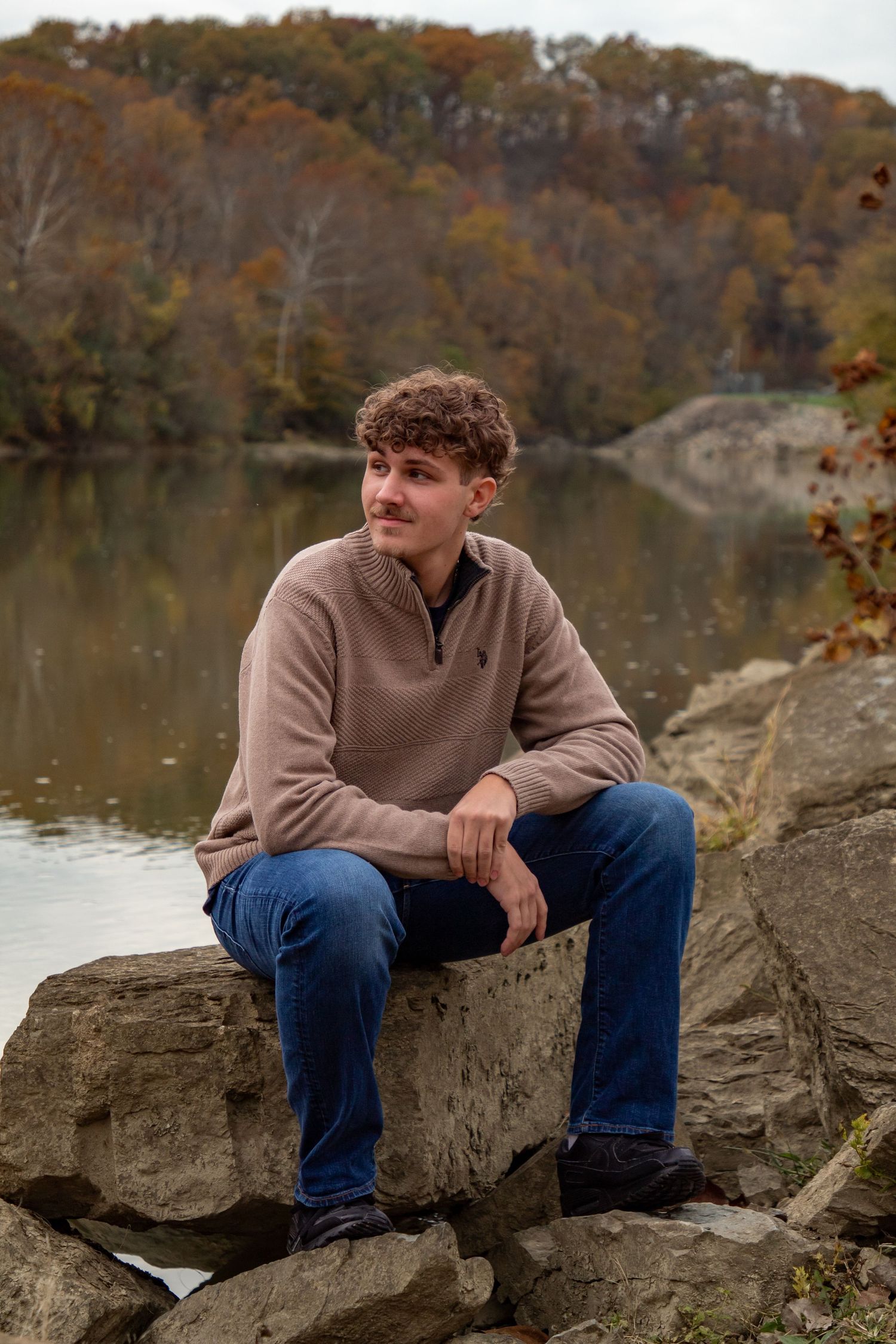 Person in beige sweater sitting on rocks by autumn lake with fall foliage in background.