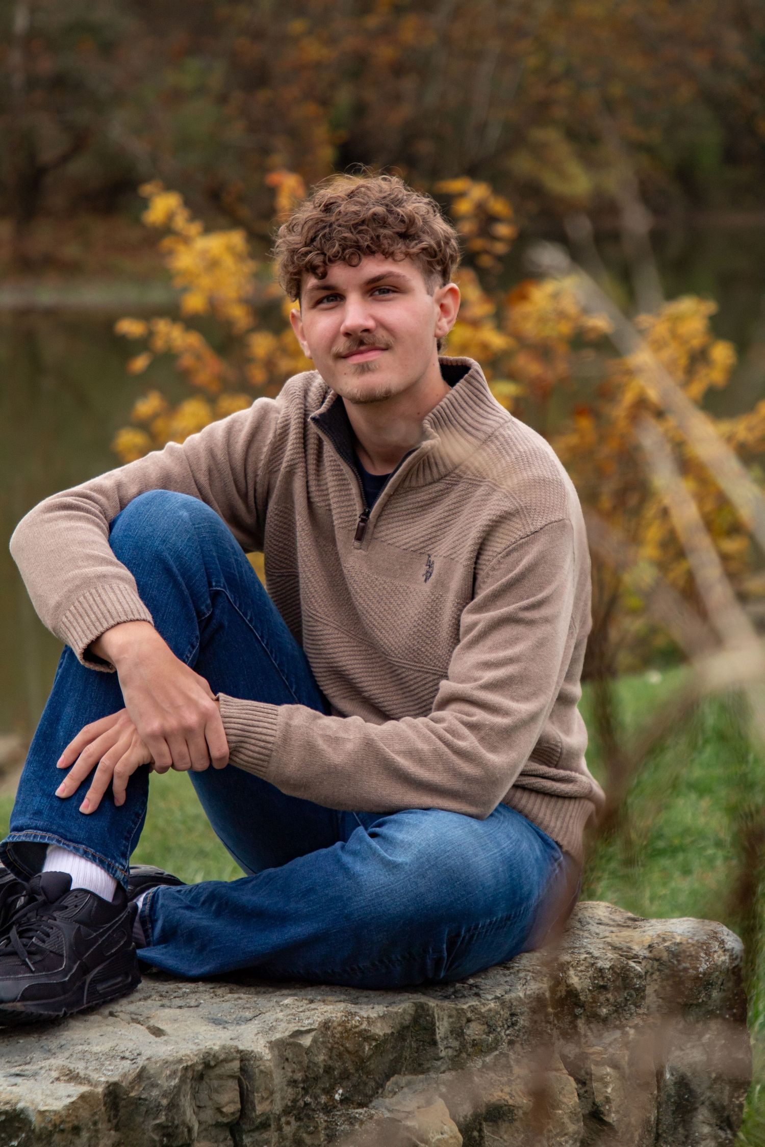 A person in a beige sweater and jeans poses casually on a stone wall during autumn with trees in the background.
