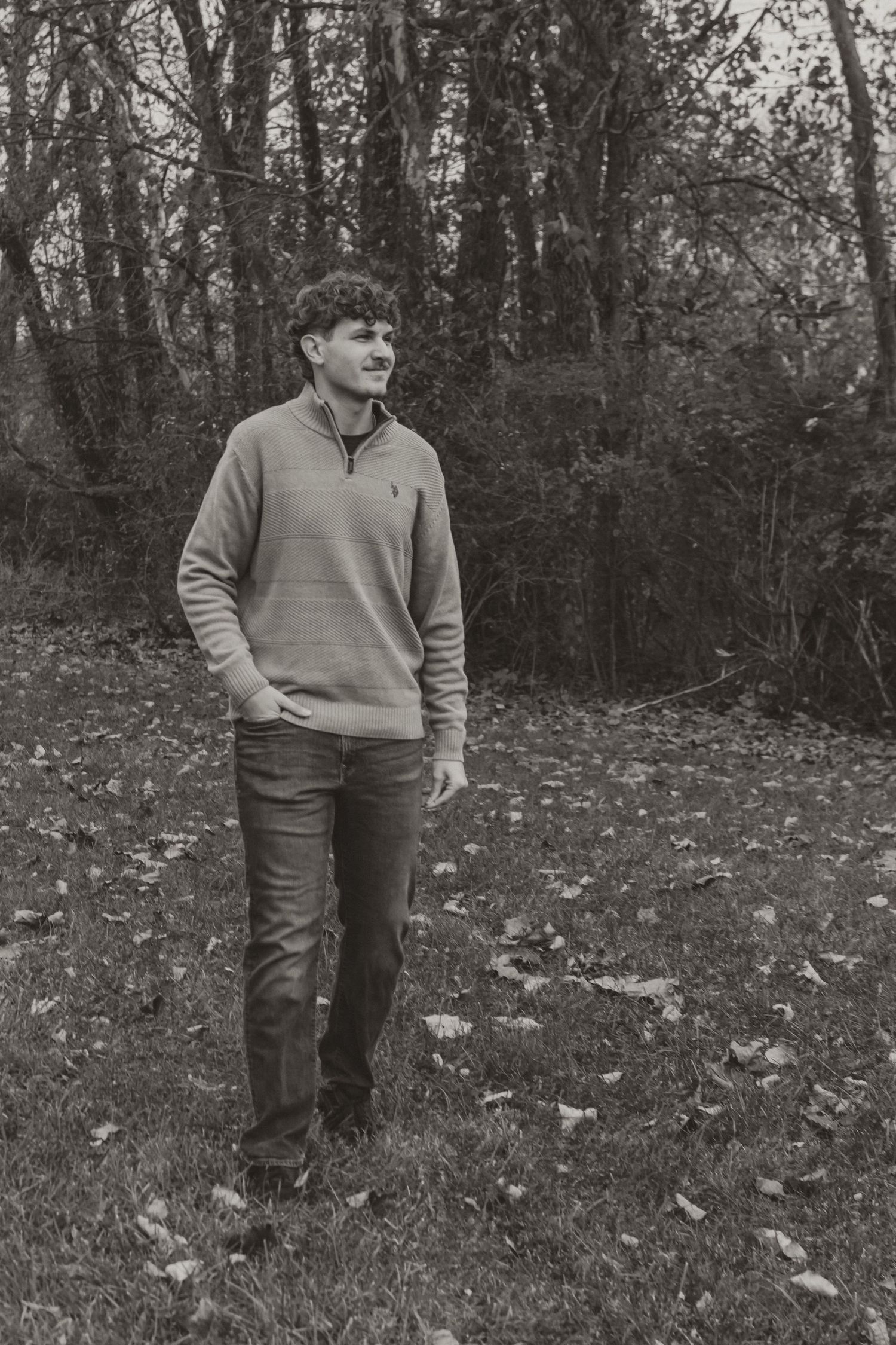 A person wearing a light colored sweater stands in a wooded area covered with fallen autumn leaves.