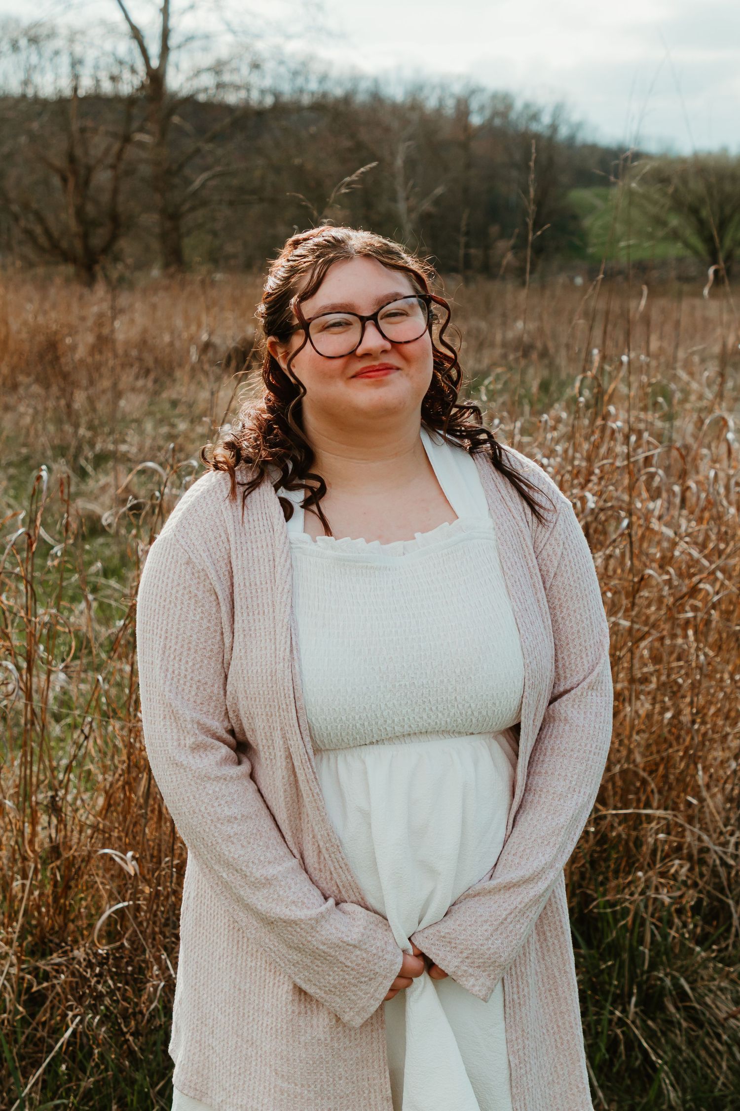 Someone in a white dress and beige cardigan stands smiling in a rustic autumn field with tall grass.