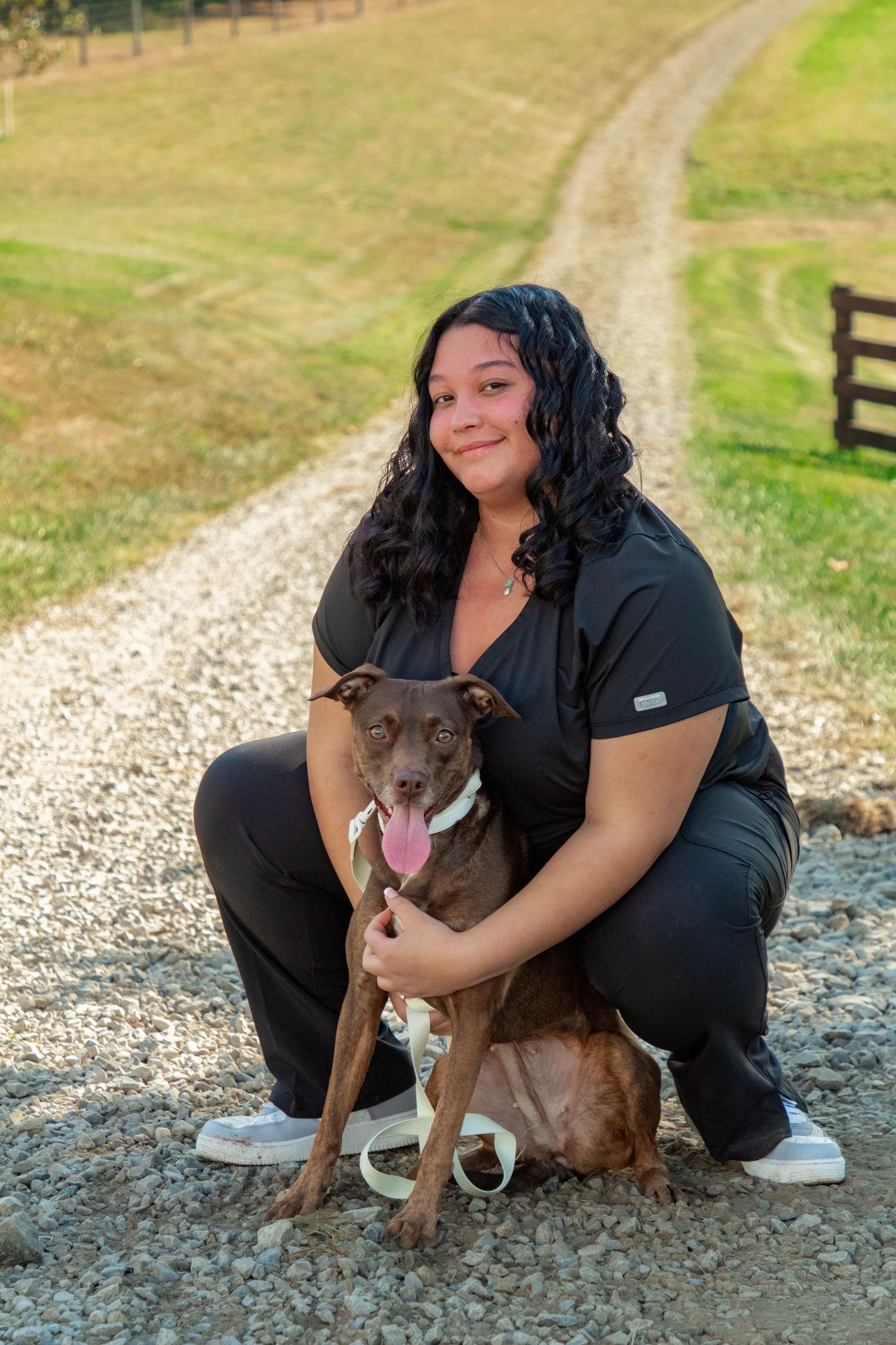 A woman in black clothing poses with a friendly brown pit bull dog on a gravel path near a grassy field.