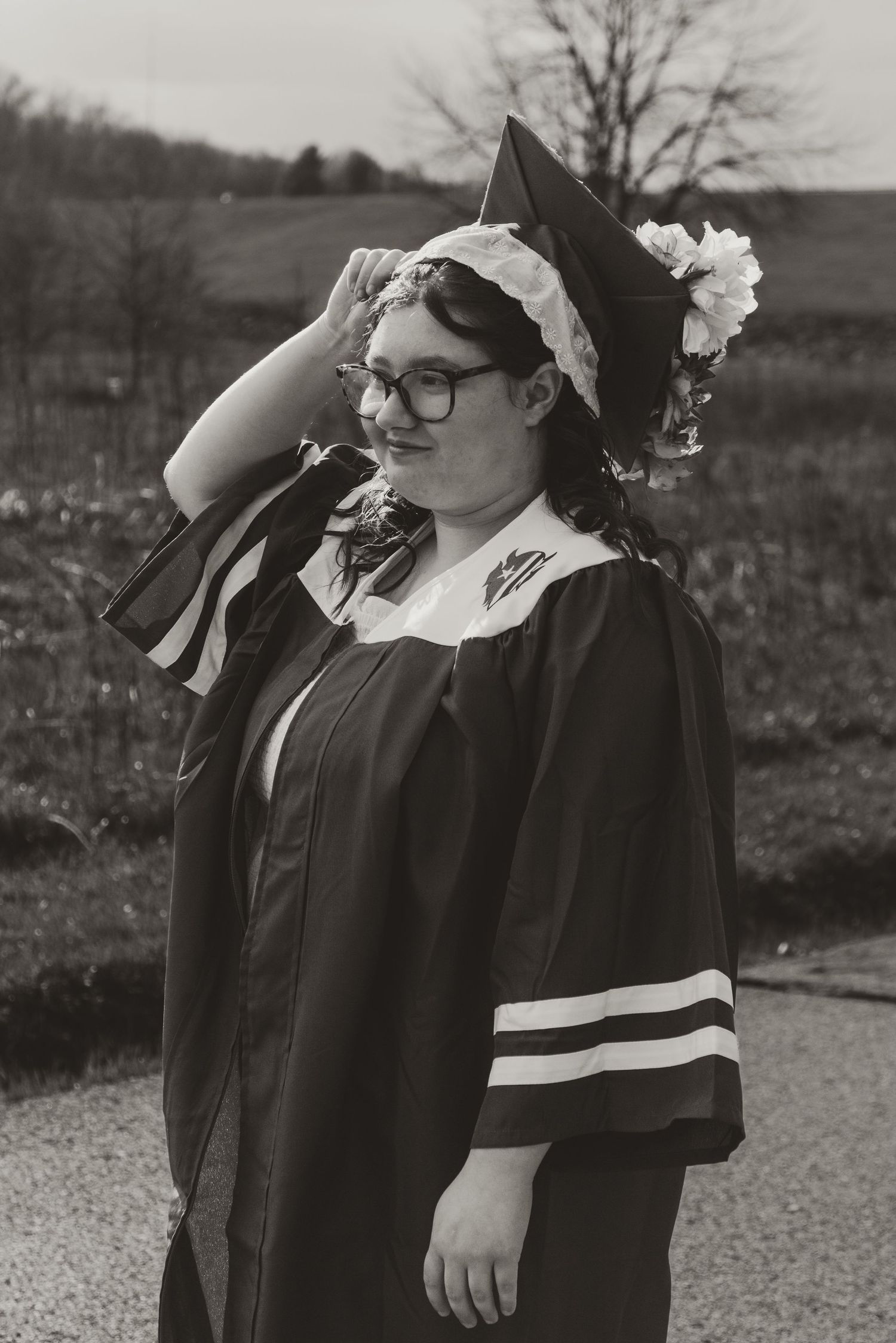 Black and white graduation photo showing graduate in academic regalia with decorated cap and glasses outdoors.