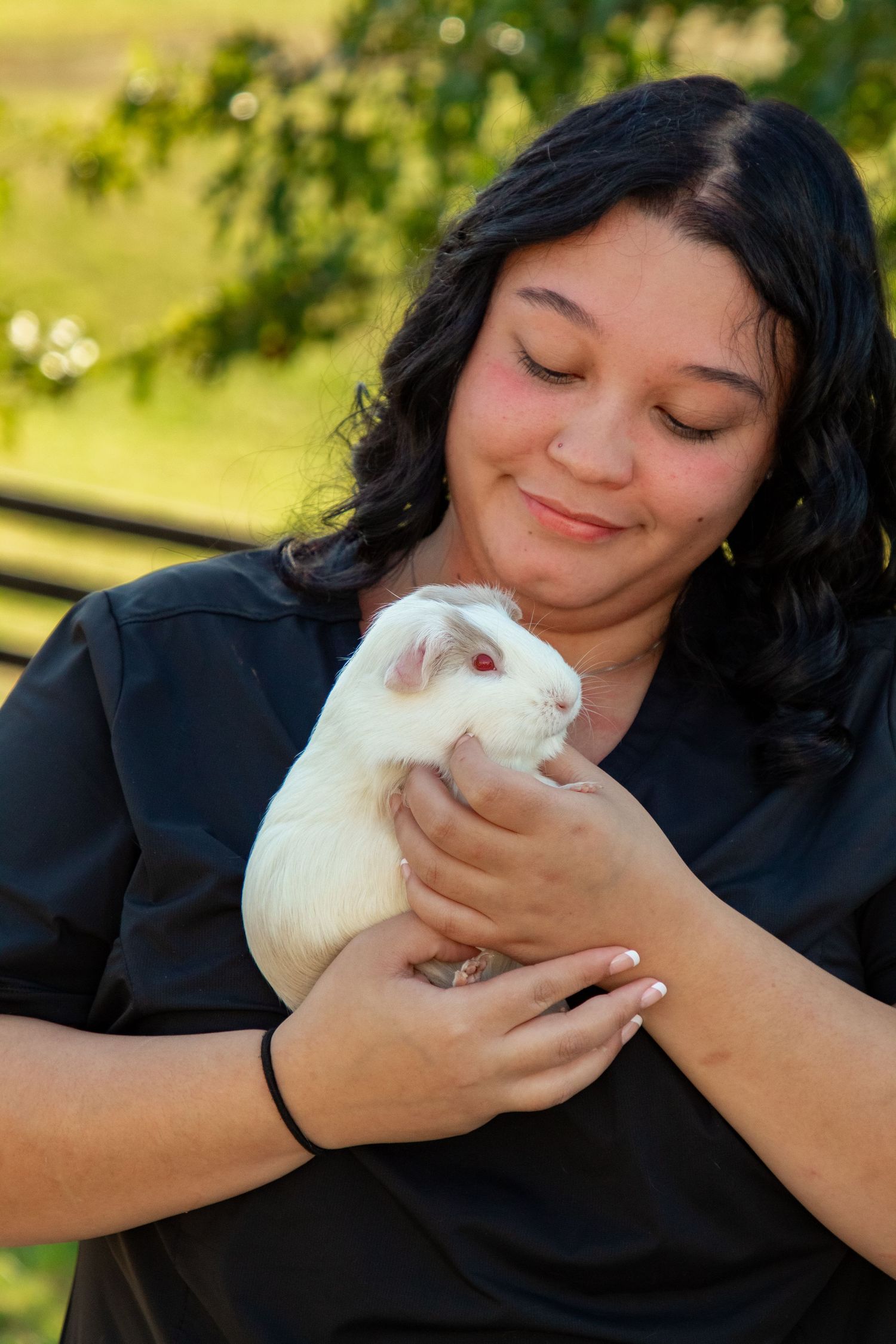 A person in black holds and cuddles with a small white bunny rabbit outdoors in a sunny garden setting.