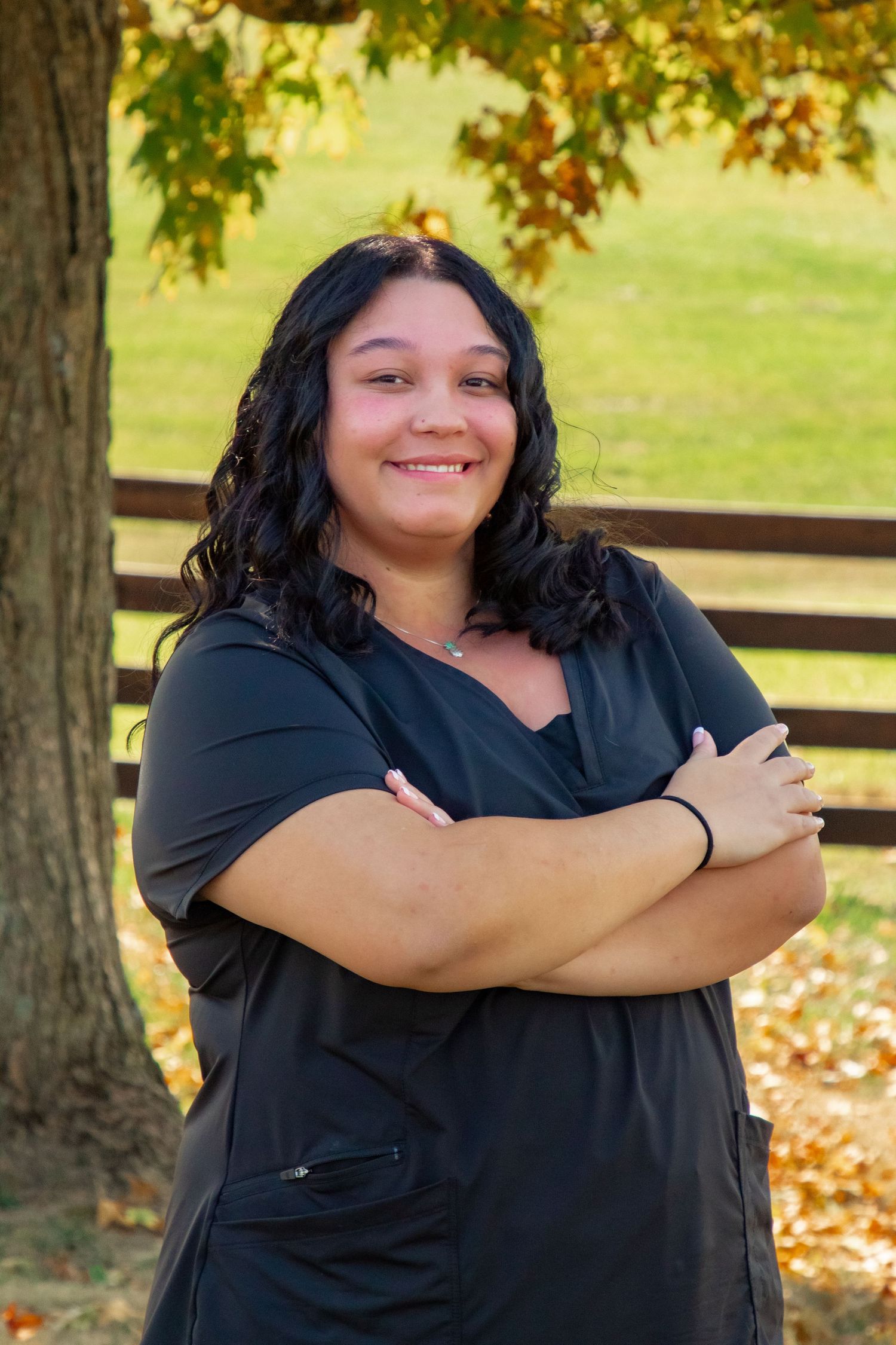A person wearing a black shirt poses with crossed arms against an autumn backdrop with trees and fallen leaves.