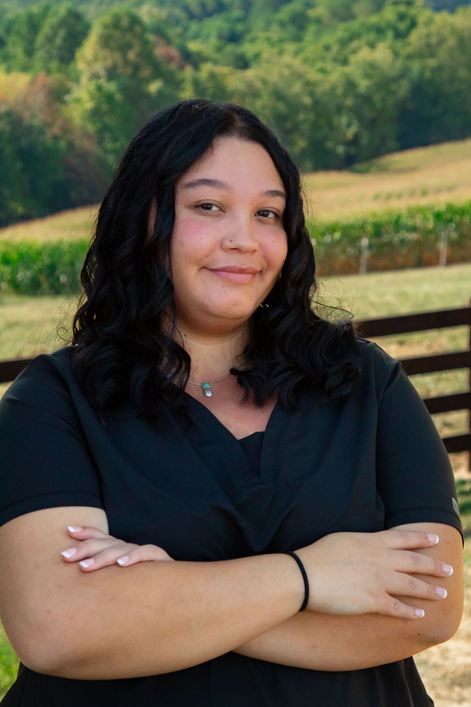 A person in dark clothing stands outdoors against a scenic rural background with rolling hills and greenery.
