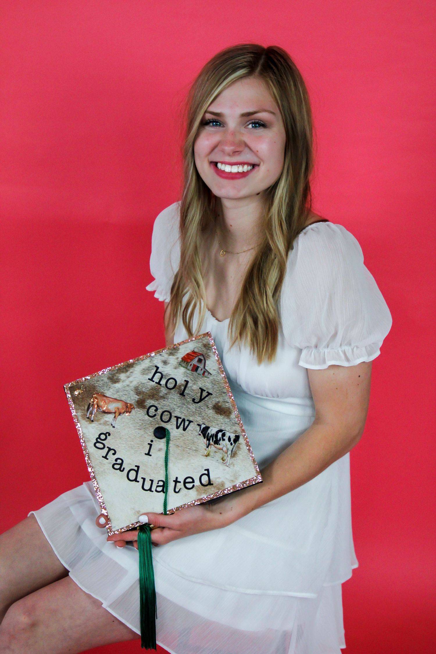 Graduate in white dress holding decorated graduation cap against pink background.