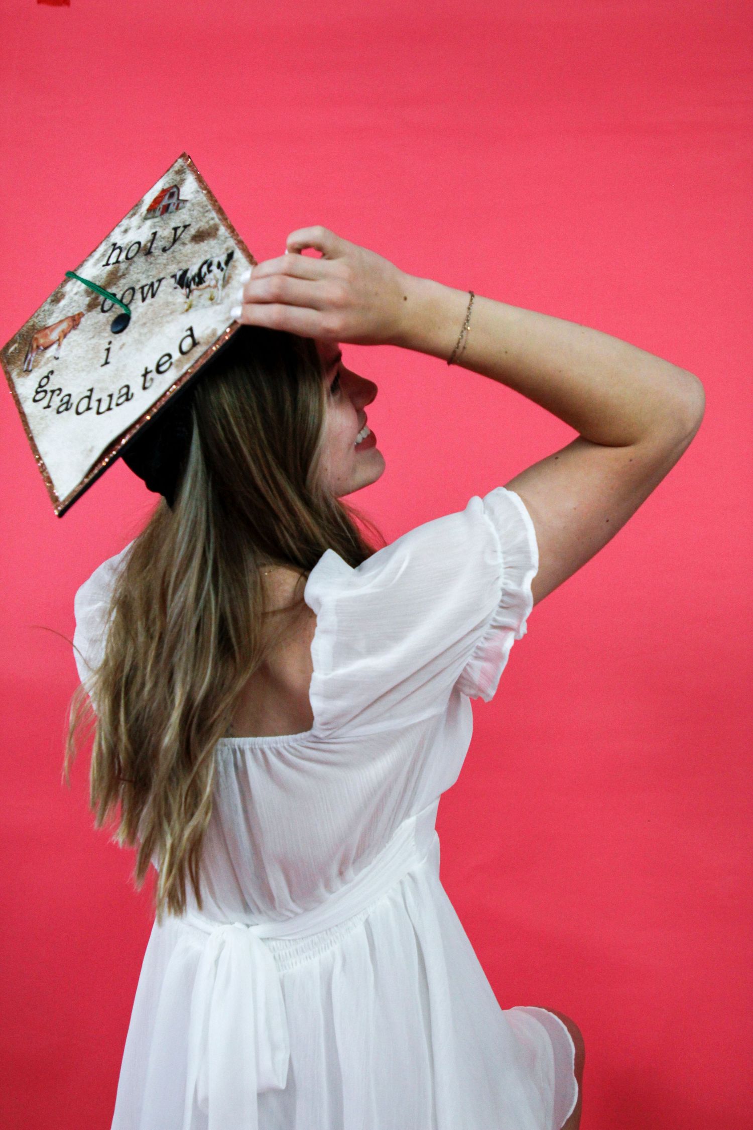 Graduate in white dress poses artistically with decorated cap against pink background.