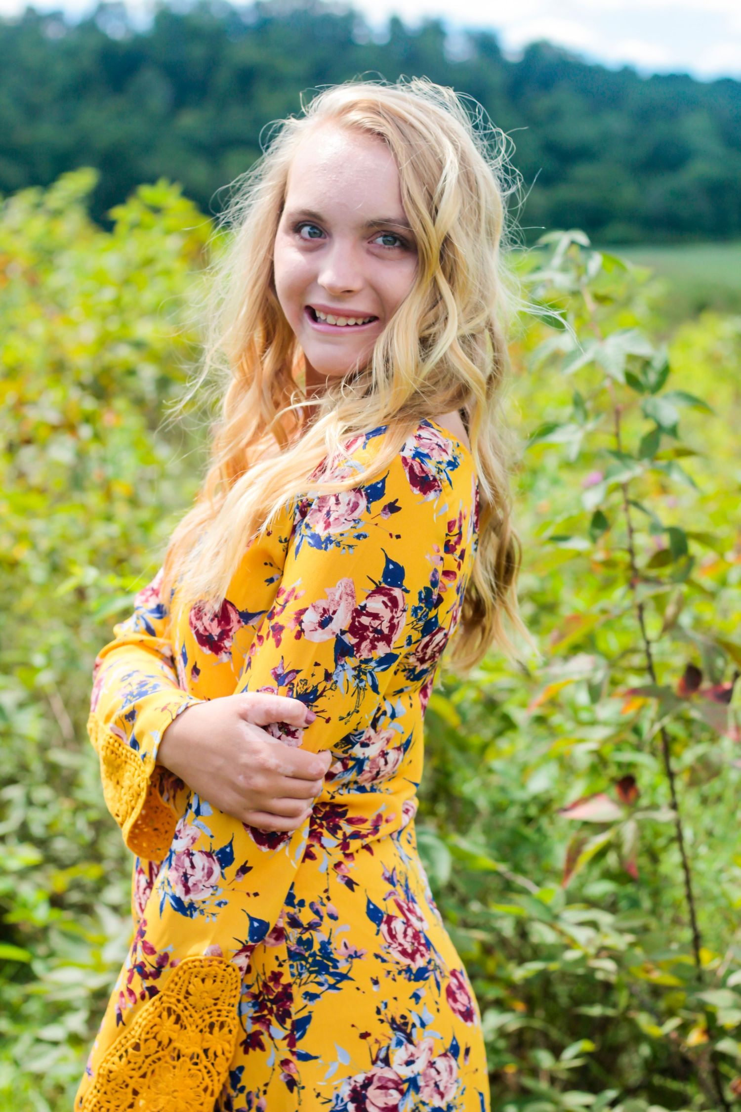 A portrait in a yellow floral dress standing in a sunny field.