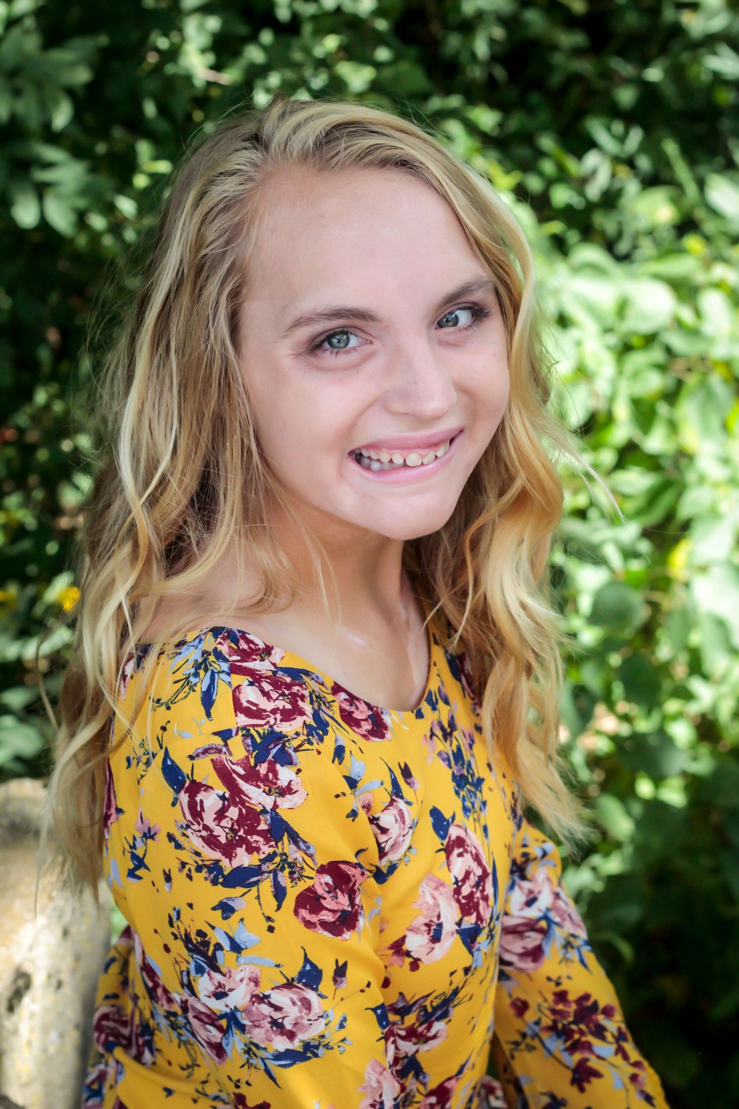 Someone in a yellow floral dress poses against green foliage.