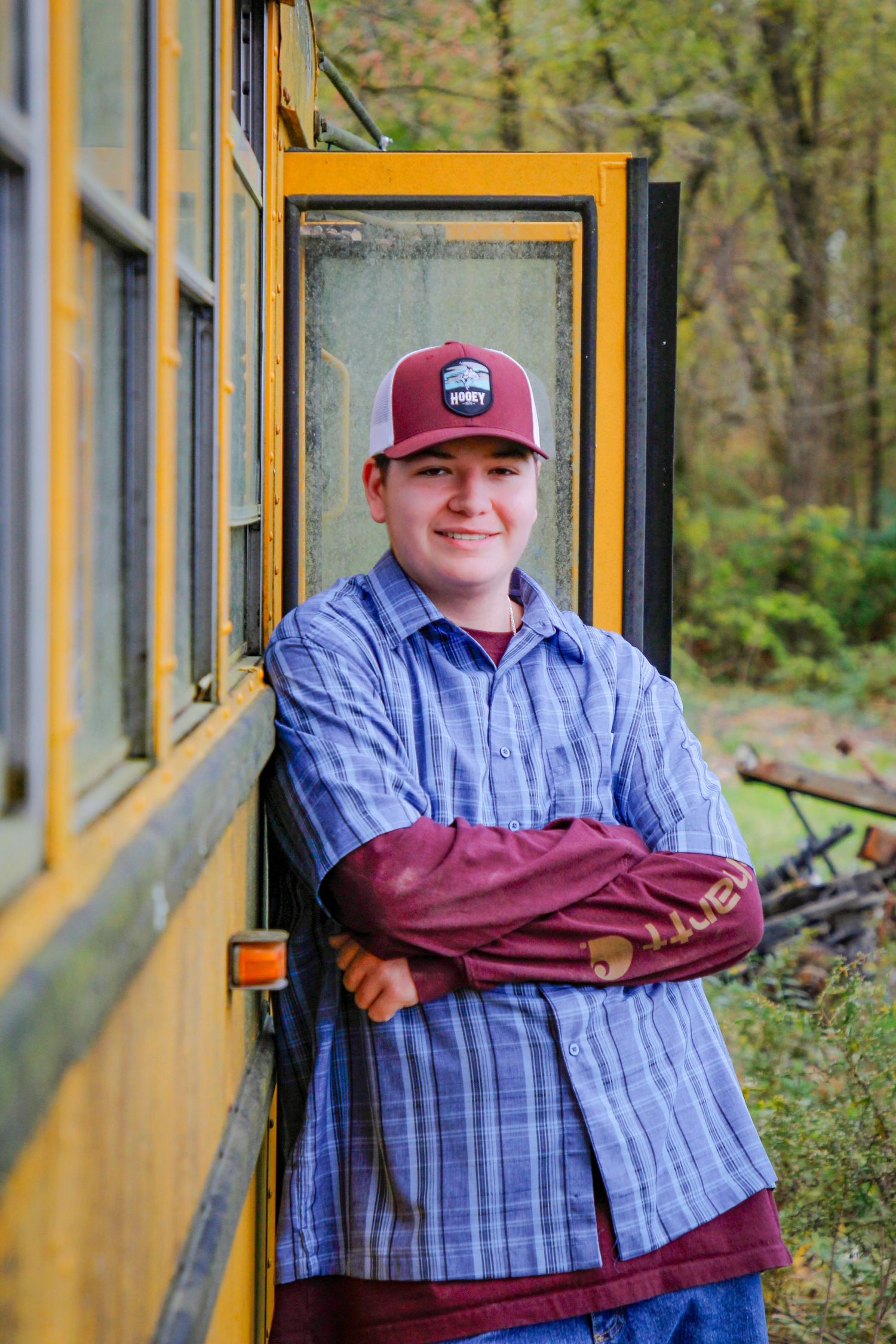 Casual pose leaning against yellow school bus wearing plaid shirt and burgundy baseball cap.
