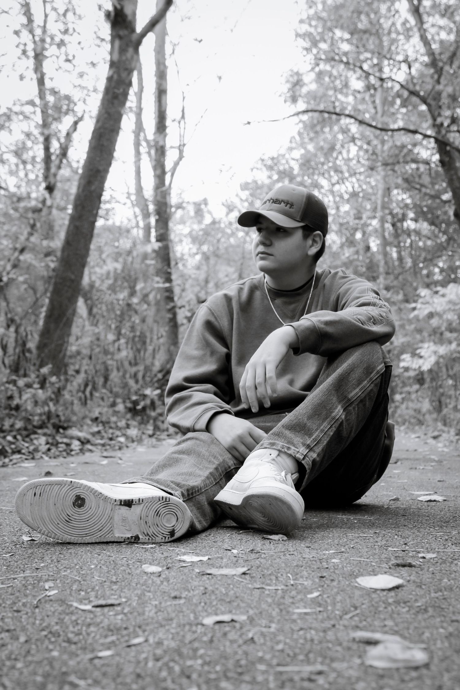 Person in casual attire and baseball cap sitting on ground in autumn forest setting.