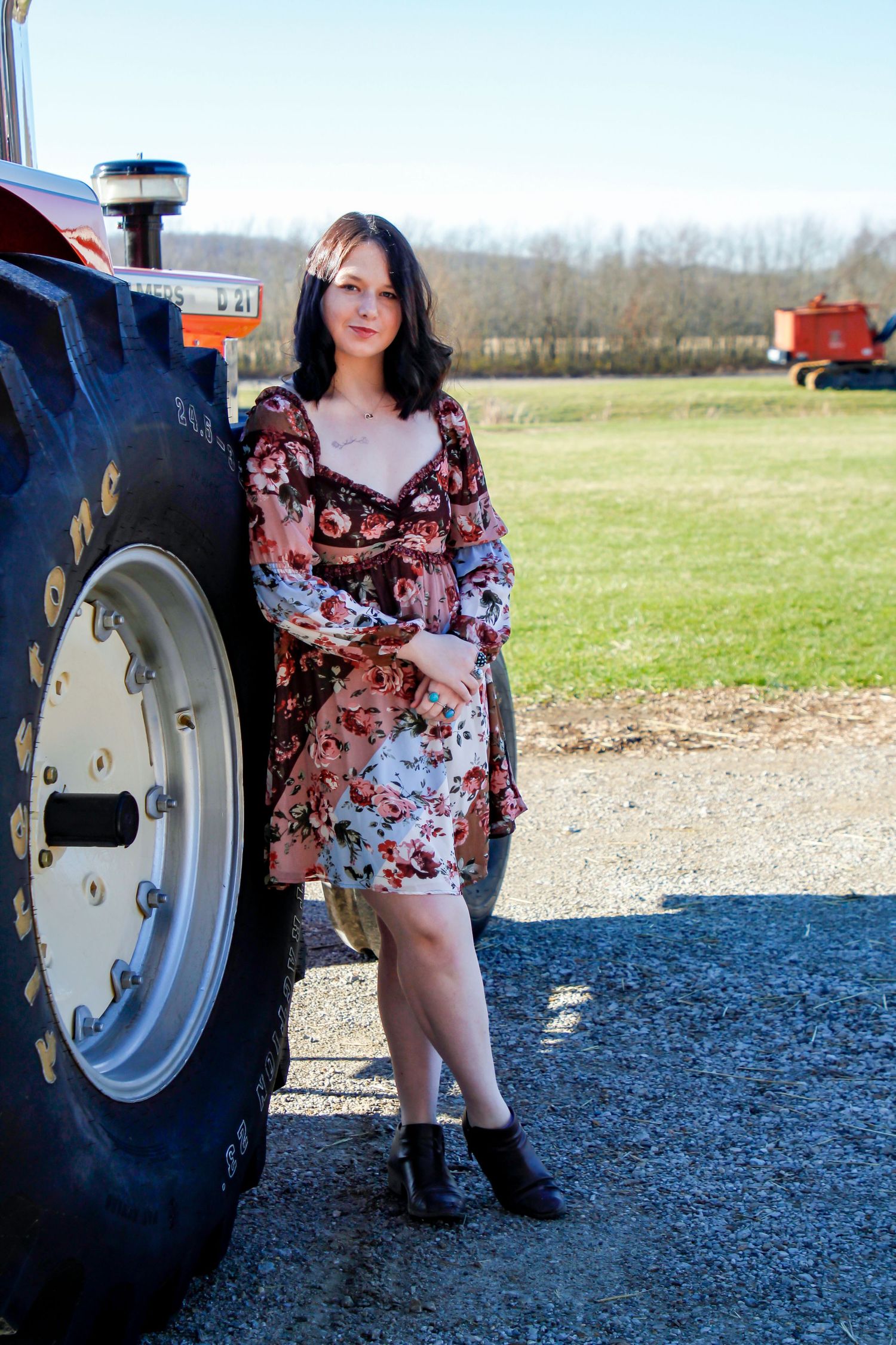 Someone in a floral dress poses beside a large red tractor in a rural setting.