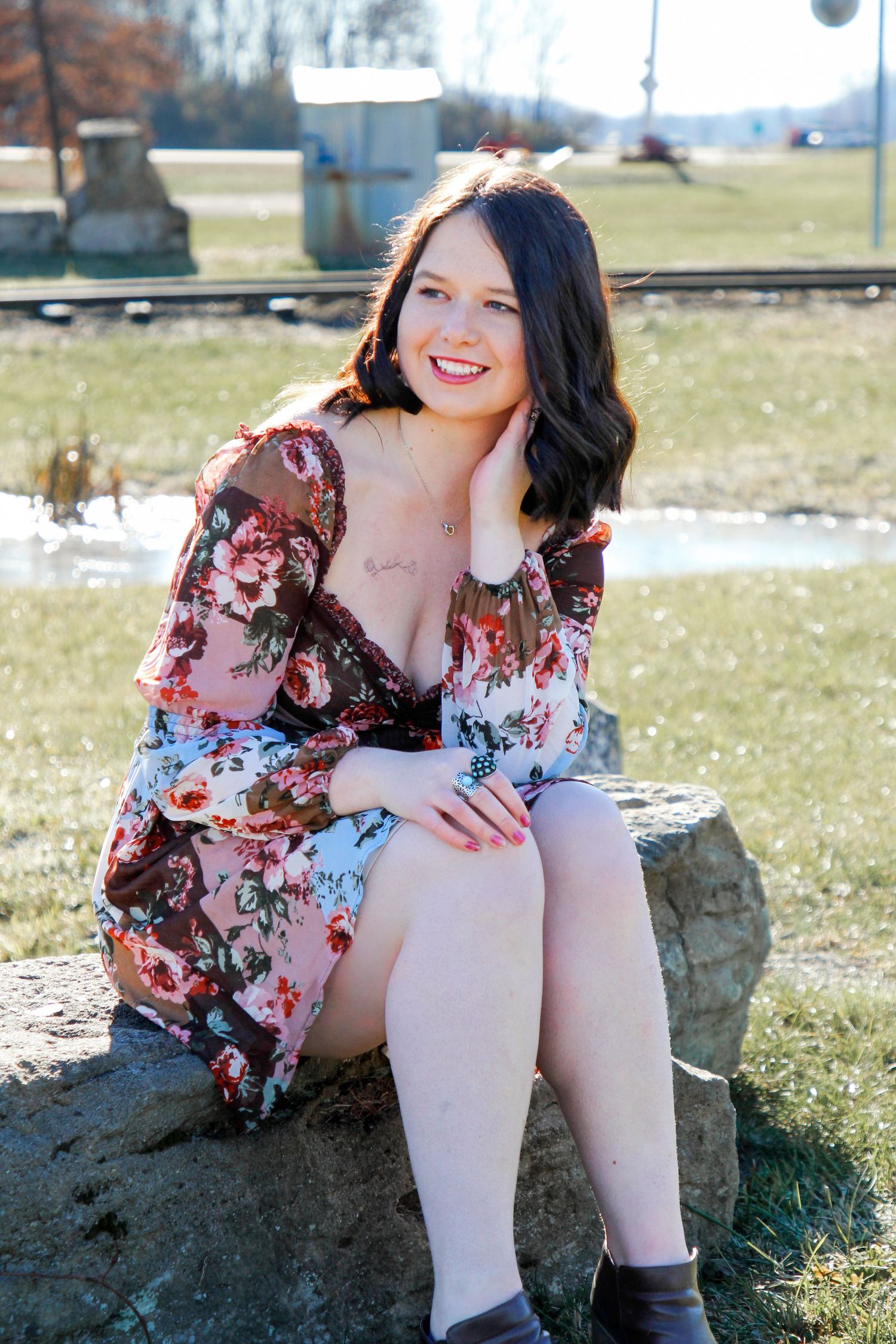 A portrait of someone in a floral dress sitting on a rock outdoors.