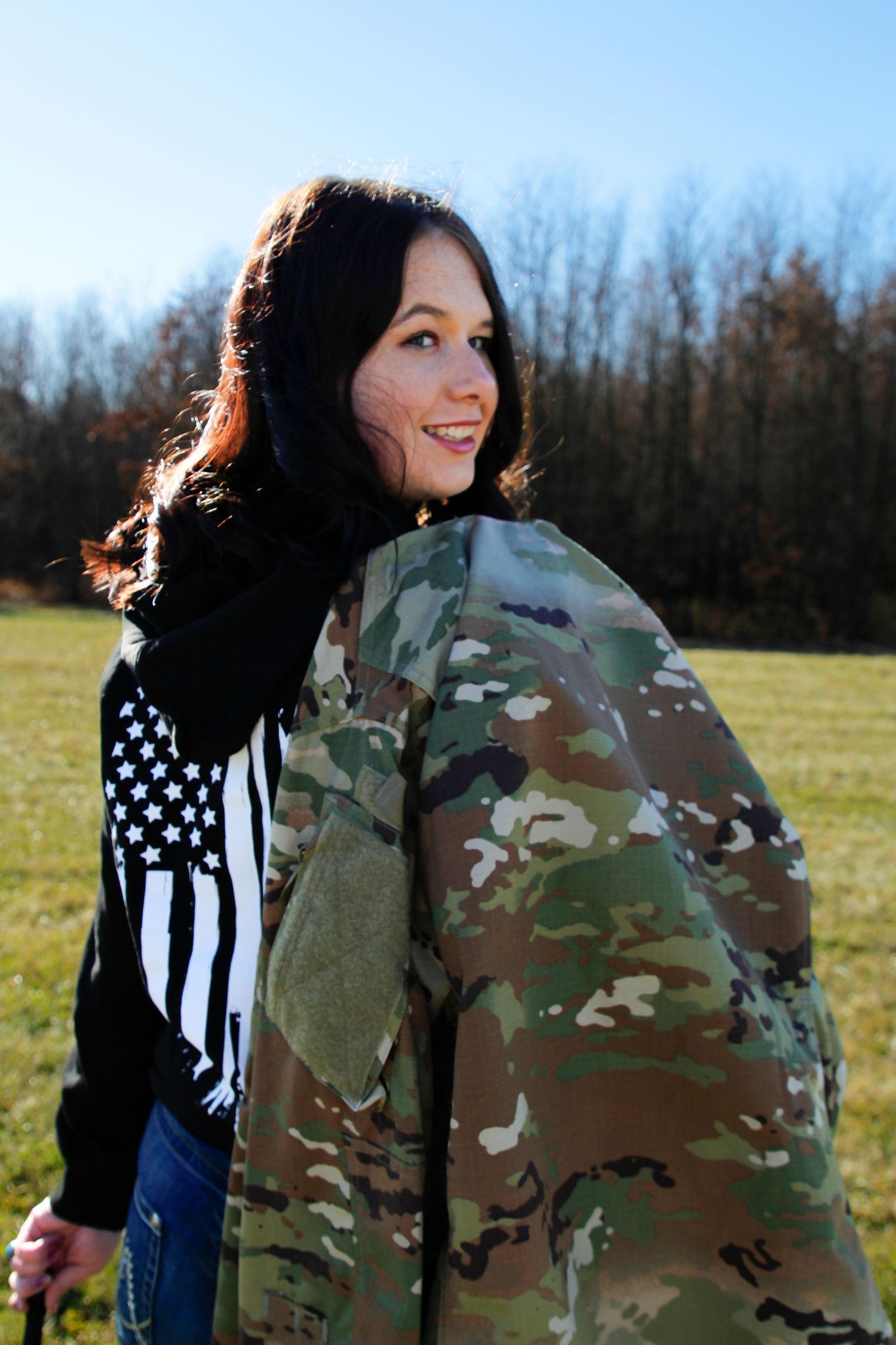 Someone poses with a camouflage jacket against a rural background.