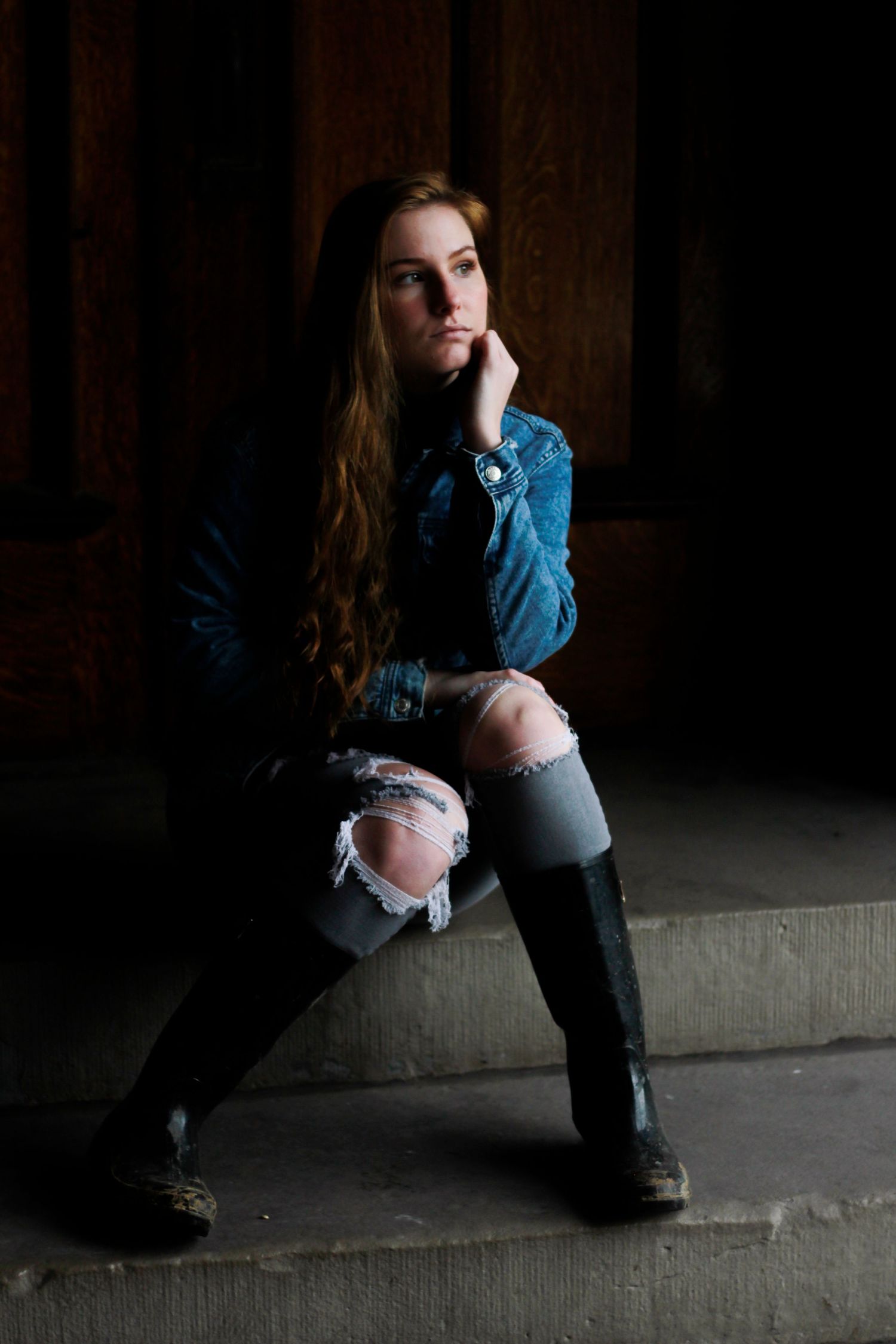 Moody portrait of figure in denim seated on concrete steps against dark wood paneling.