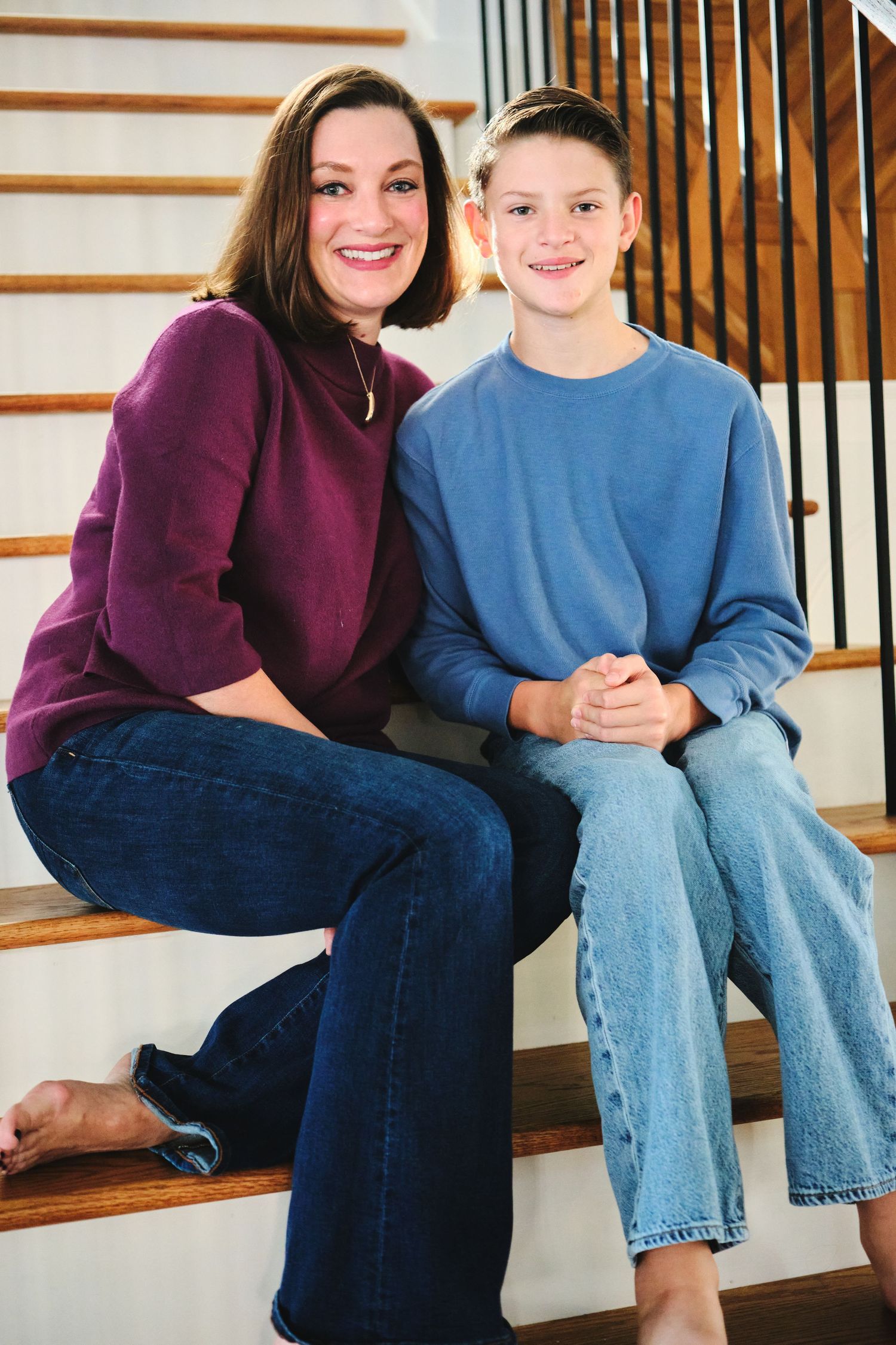A smiling woman in purple top and jeans poses with a young boy on wooden staircase at home.