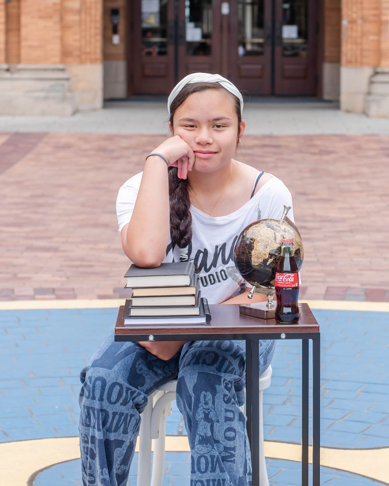 Student sits at small table with books and soda bottle in front of brick building.