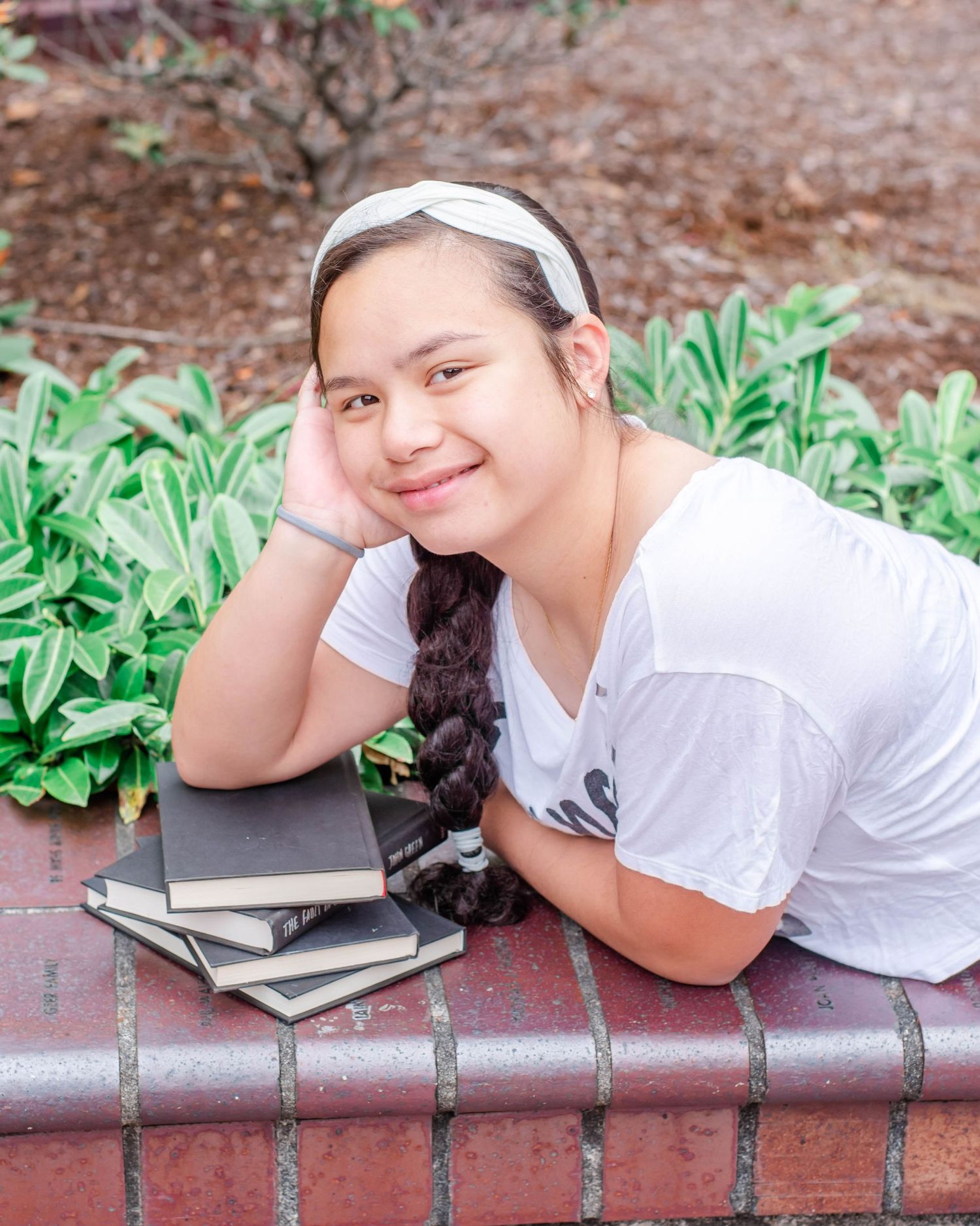 Student in white shirt lies on brick wall with stack of books nearby.