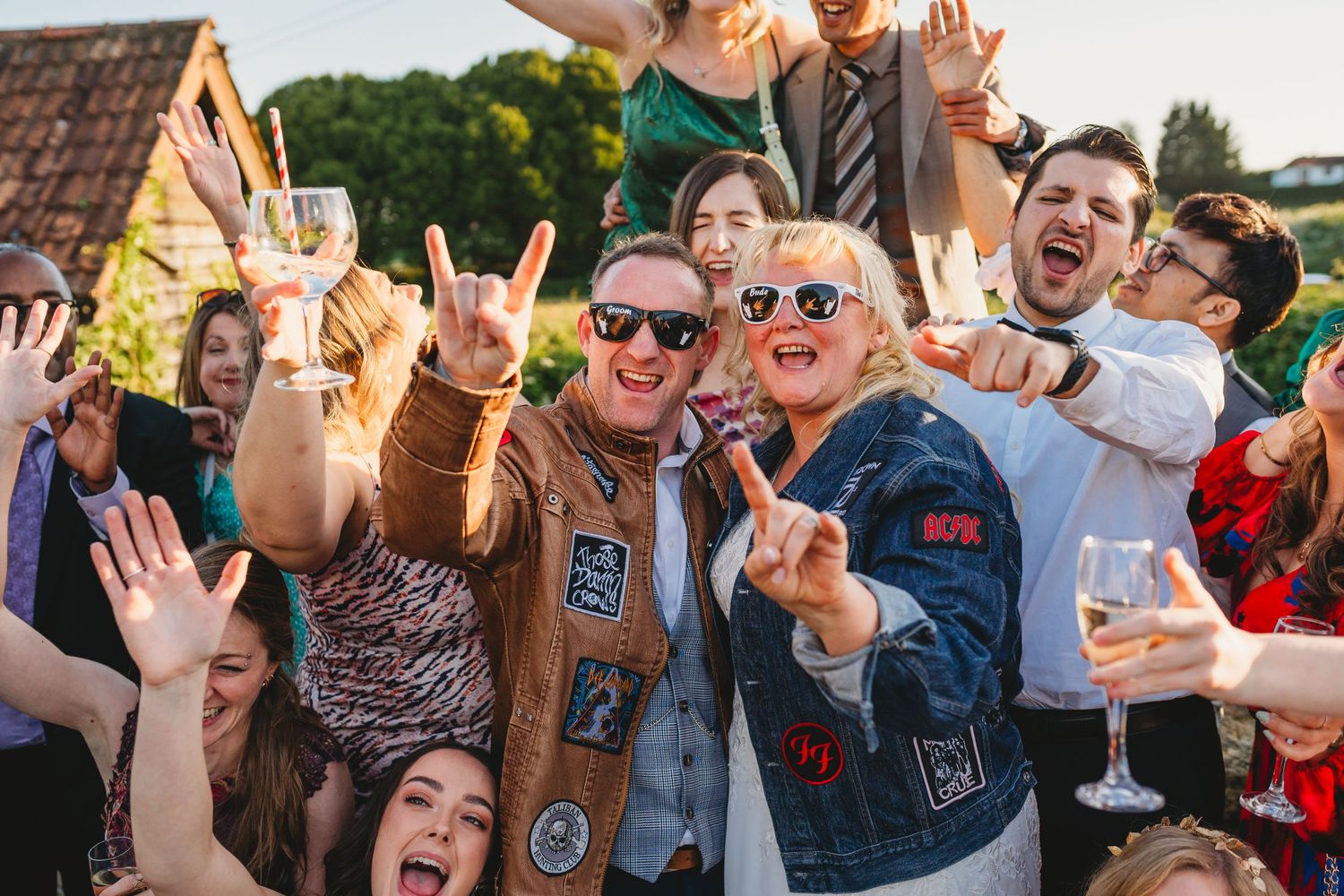 Large group of wedding guests celebrating outdoors with energetic poses and gestures.