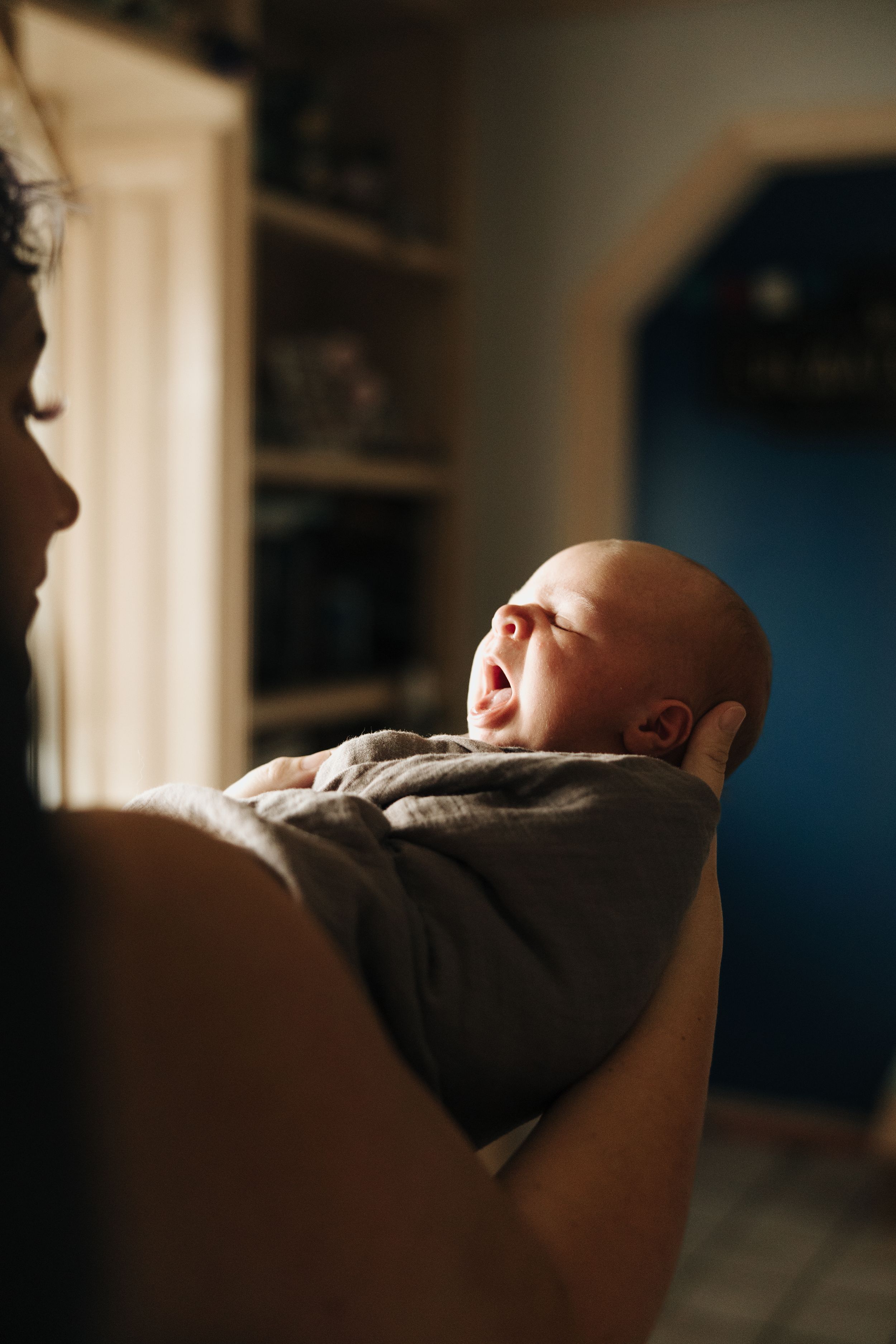 A sleeping newborn baby in grey clothing rests against someone's shoulder in a dimly lit room.