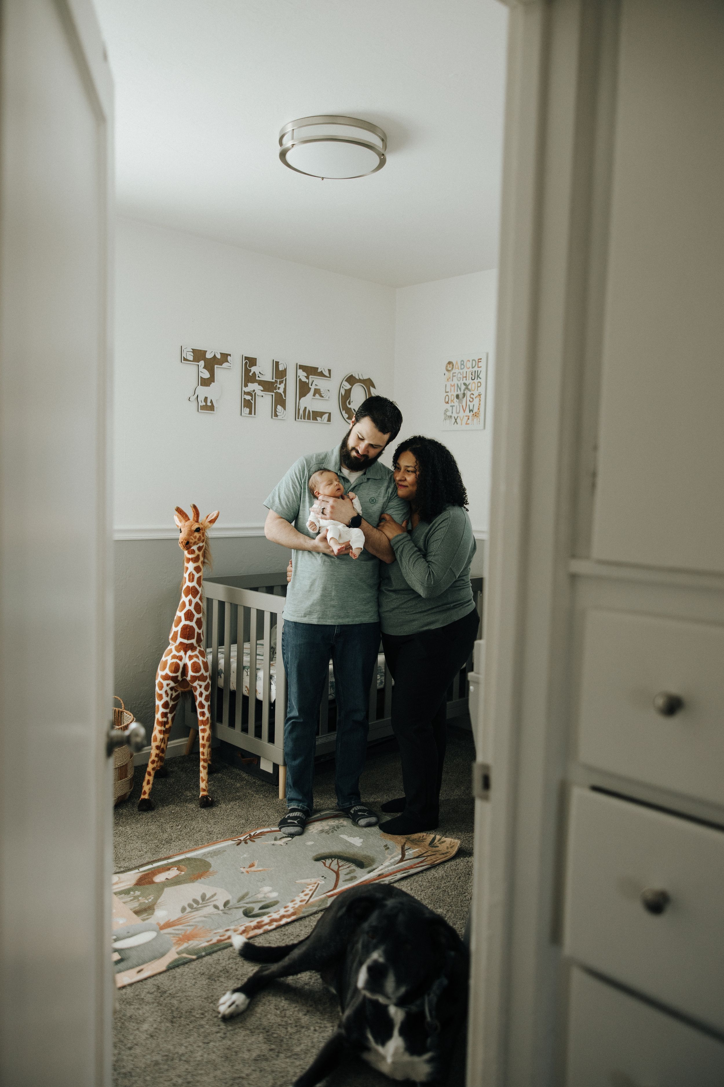 A cozy nursery with a giraffe decoration, white furniture, a dog resting on the floor, and three people by the crib.