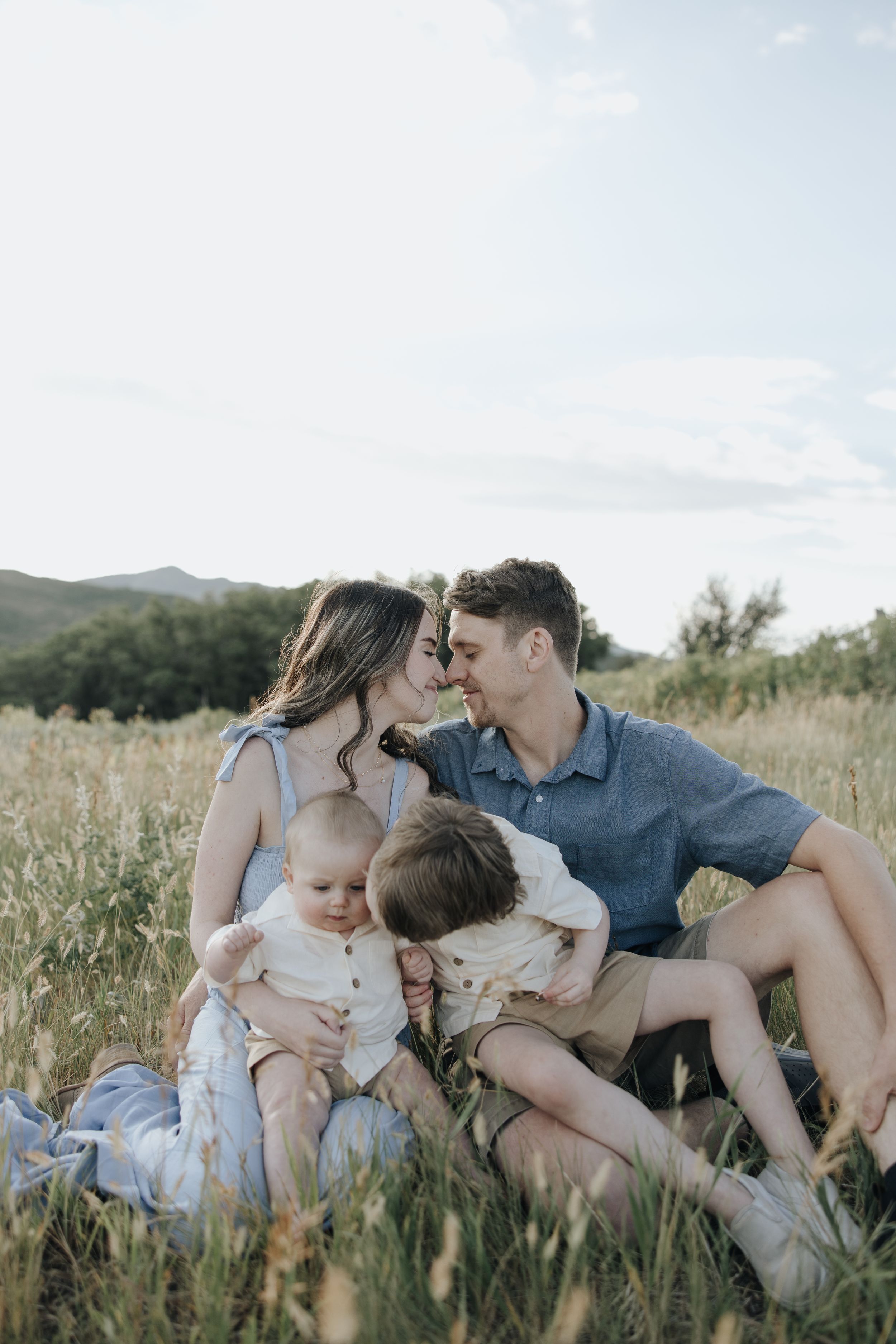 Family sits together in a golden field at sunset sharing an intimate moment among wildflowers and mountains.