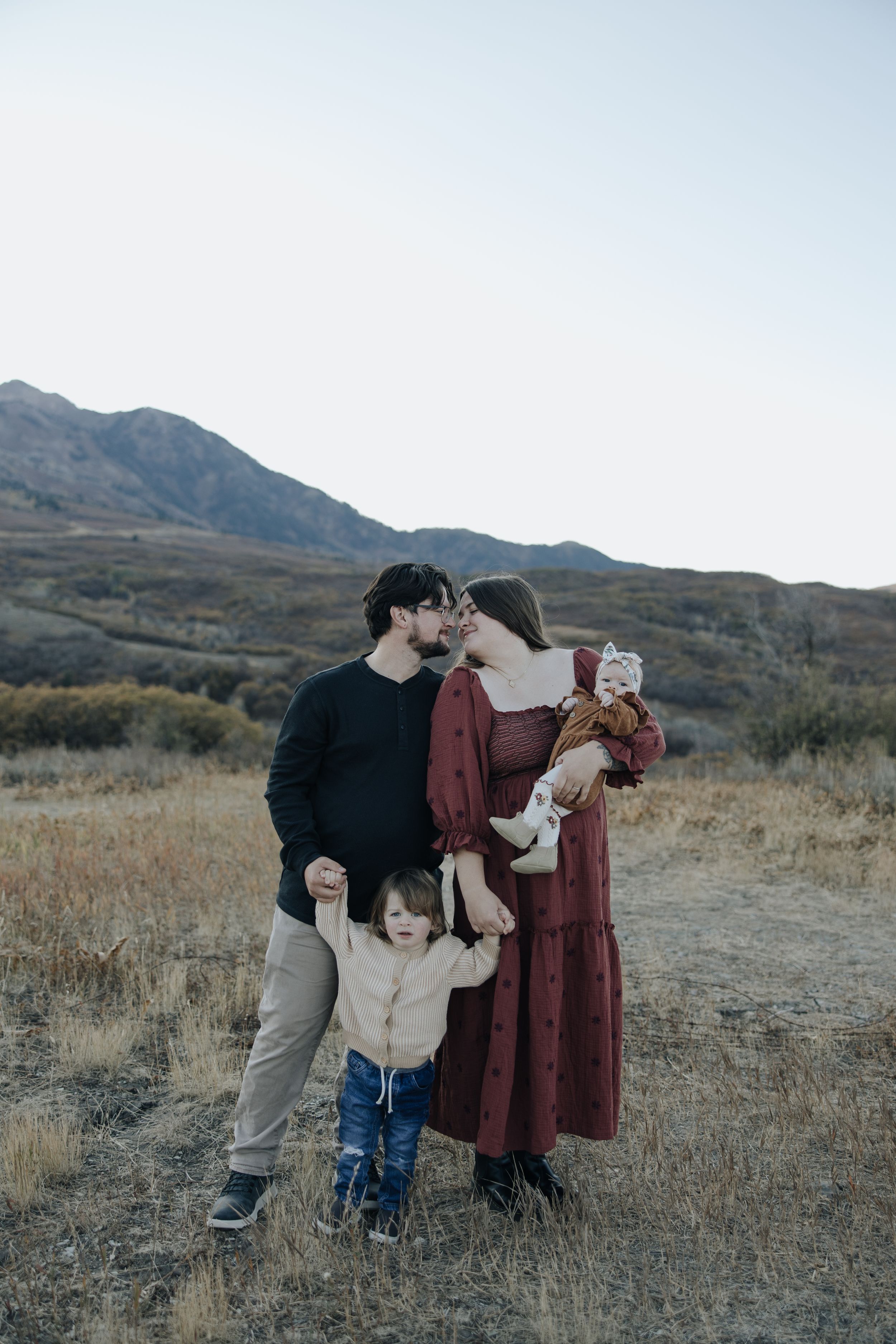 A family embraces together in an outdoor setting with mountains and rustic terrain in the background on an autumn day.
