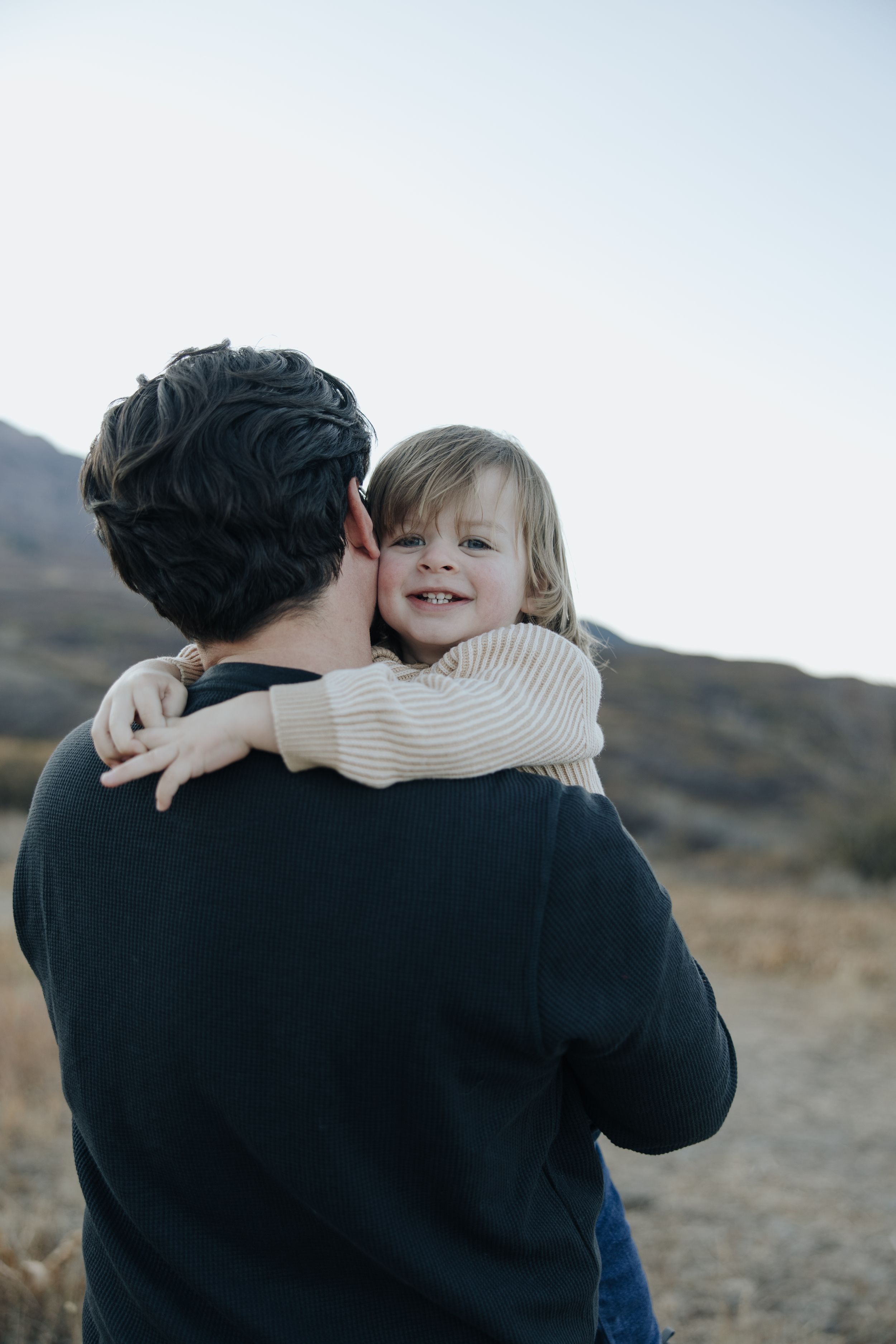 A small child in a cream sweater hugs an adult wearing dark clothing while outdoors in a mountain setting.