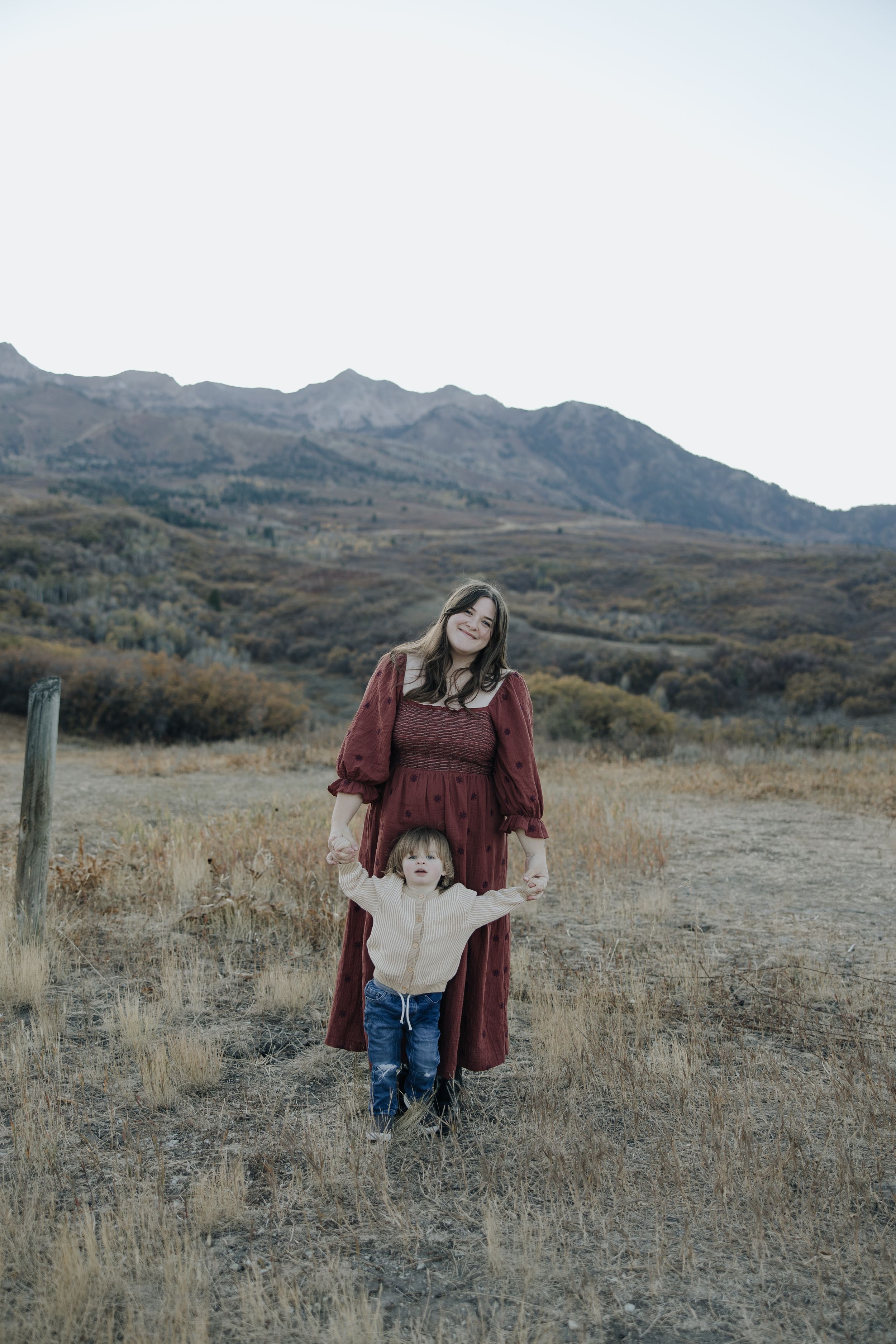 A parent and child stand together in a brown field with rugged mountains rising in the background at dusk.