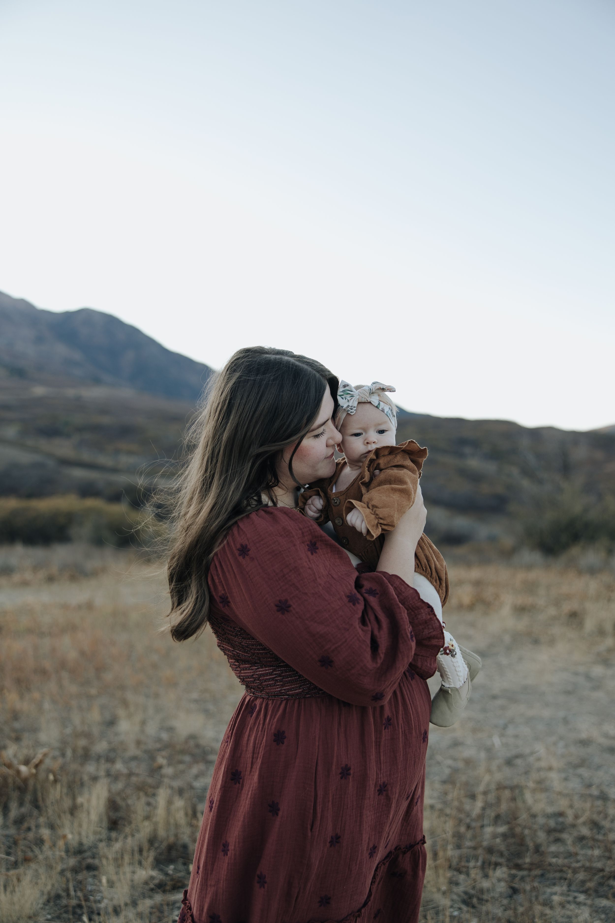 A person in a burgundy dress holds up a small fluffy cat against a mountain landscape at dusk.