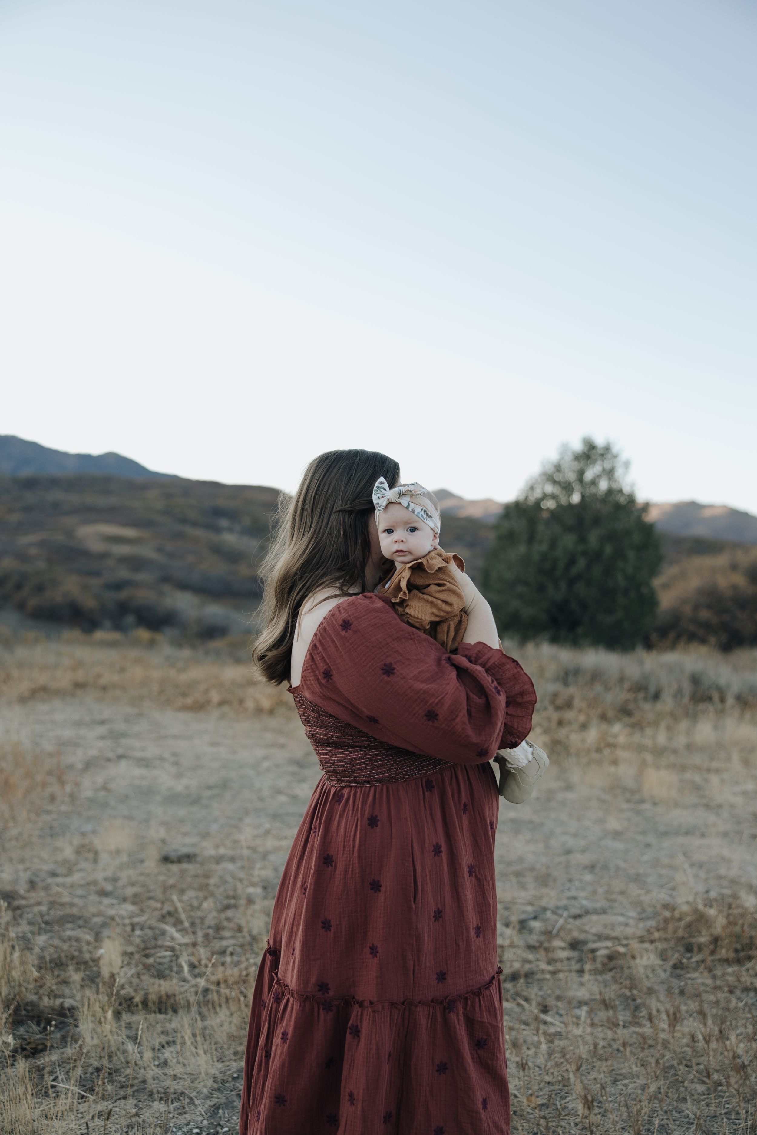 A person in a burgundy dress stands in a golden field with mountains in the background during sunset.