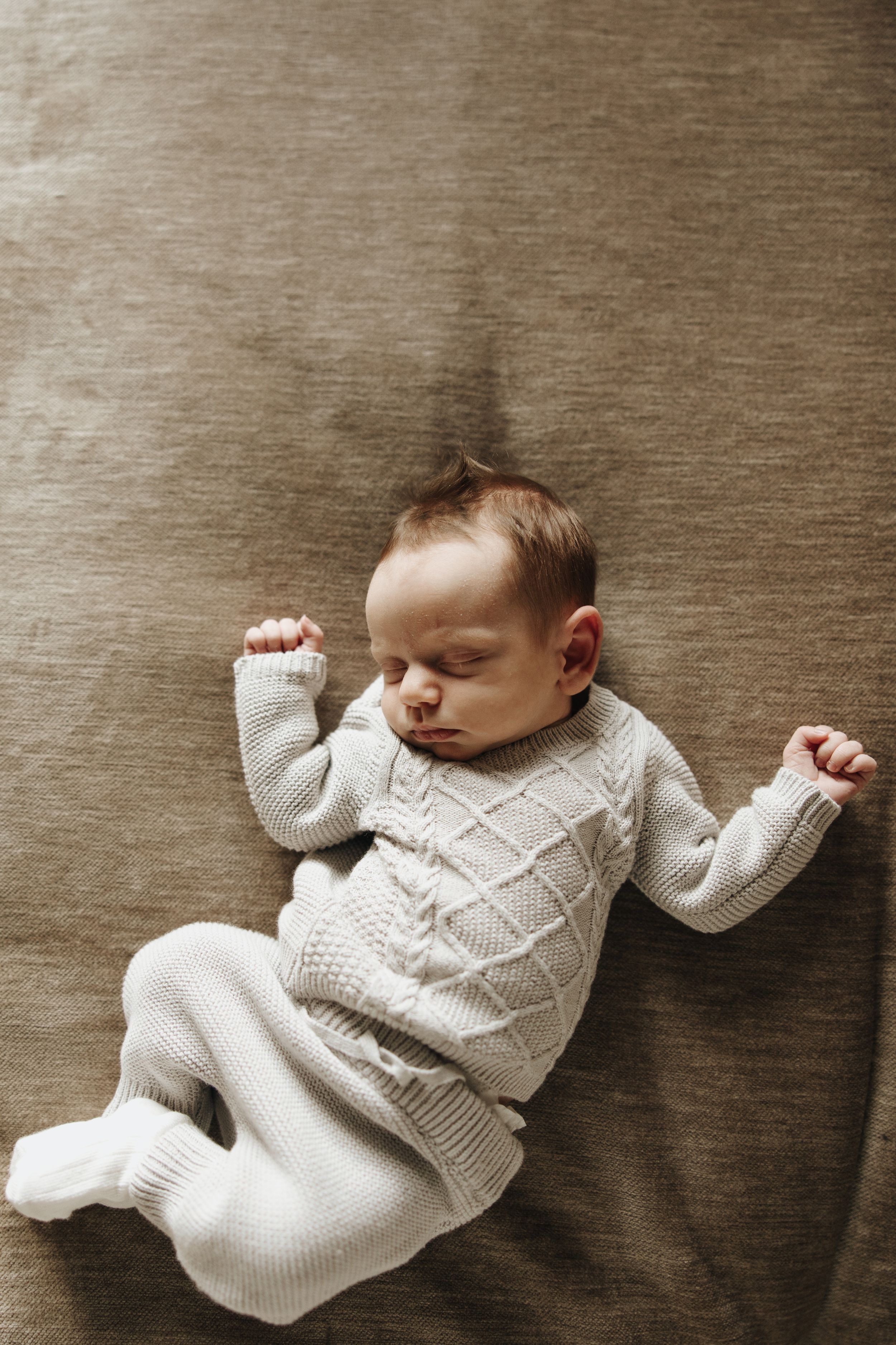 Sleeping baby wearing cozy white knit sweater and pants lying on brown textured background.