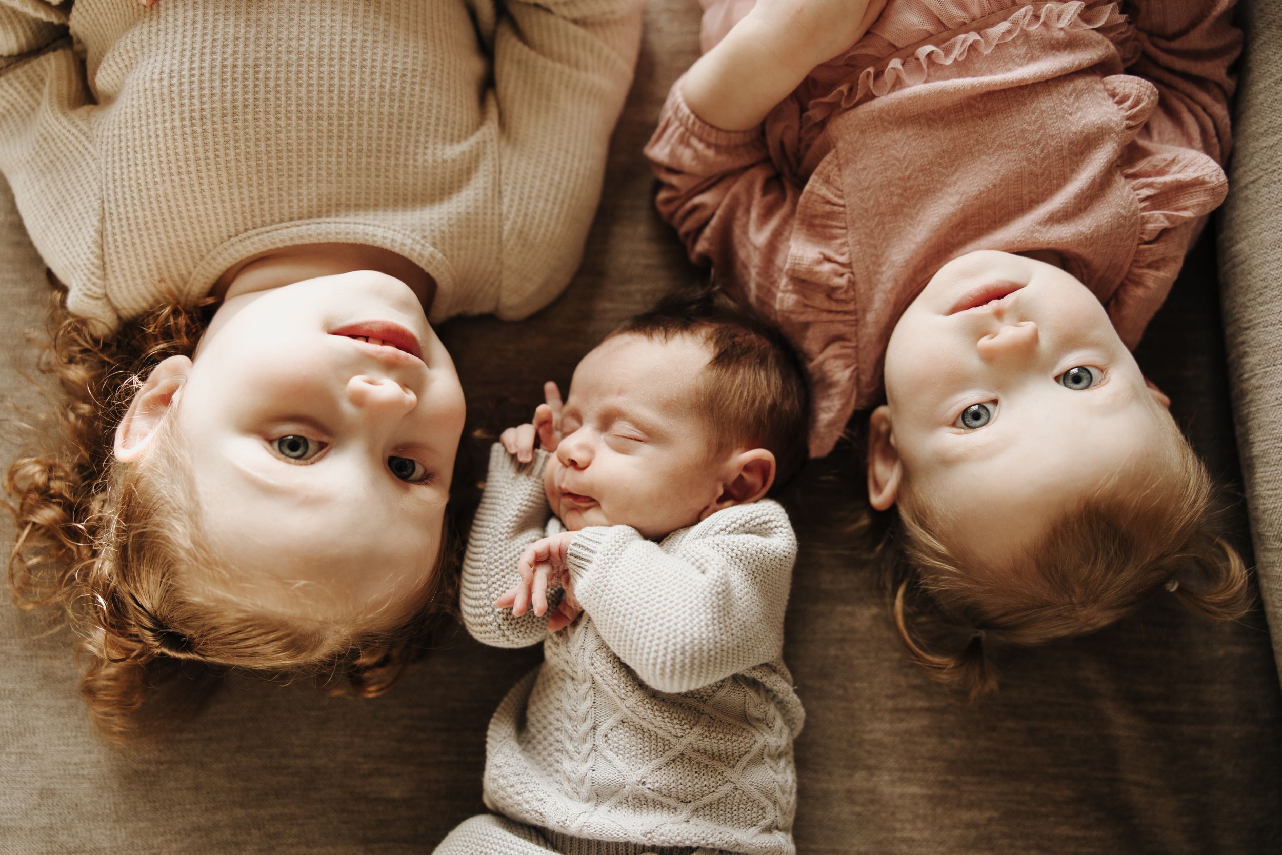Overhead view of three siblings in warm toned sweaters lying on wooden floor and looking up at camera.