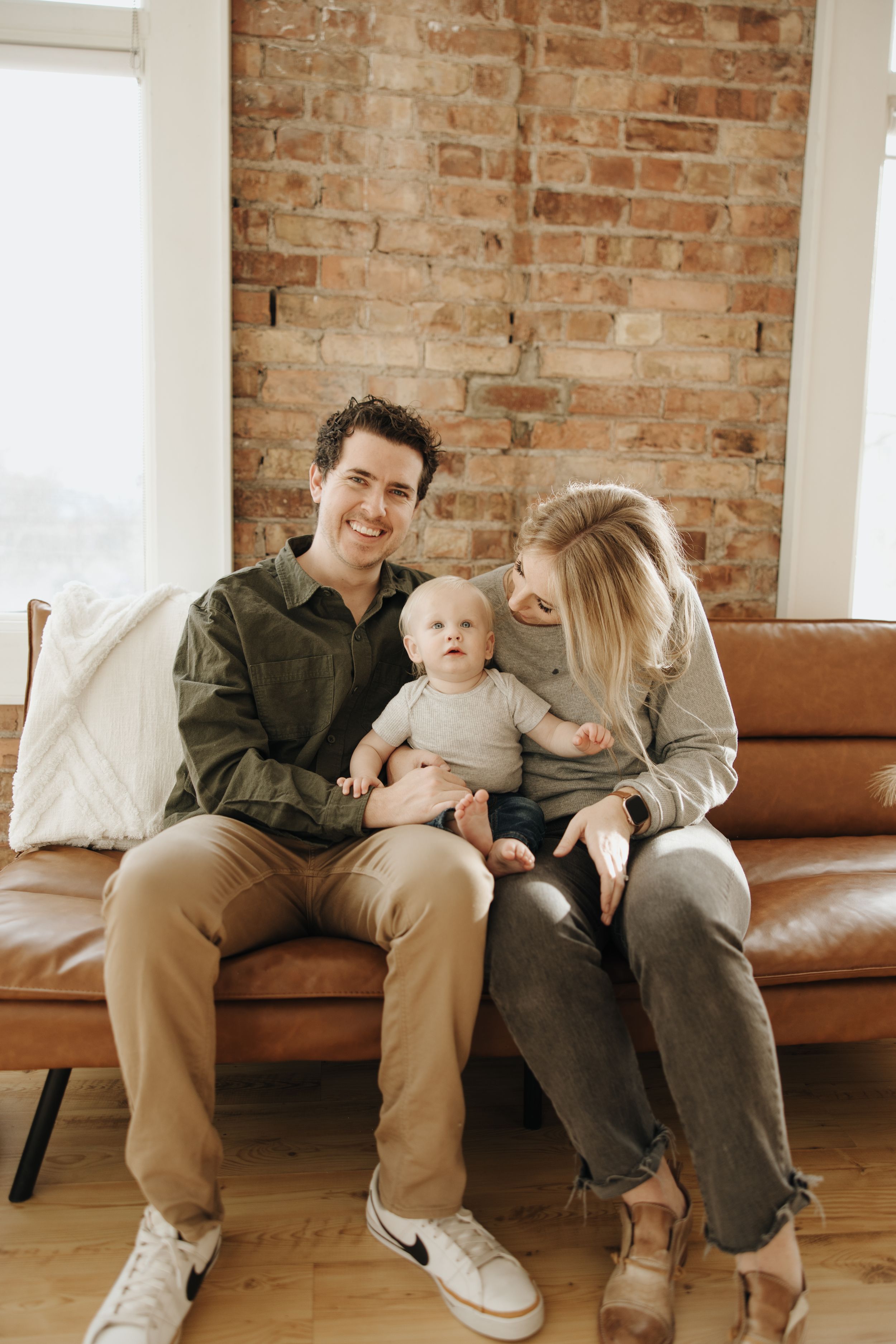 A family sits together on a brown leather couch against an exposed brick wall in a cozy living room setting.