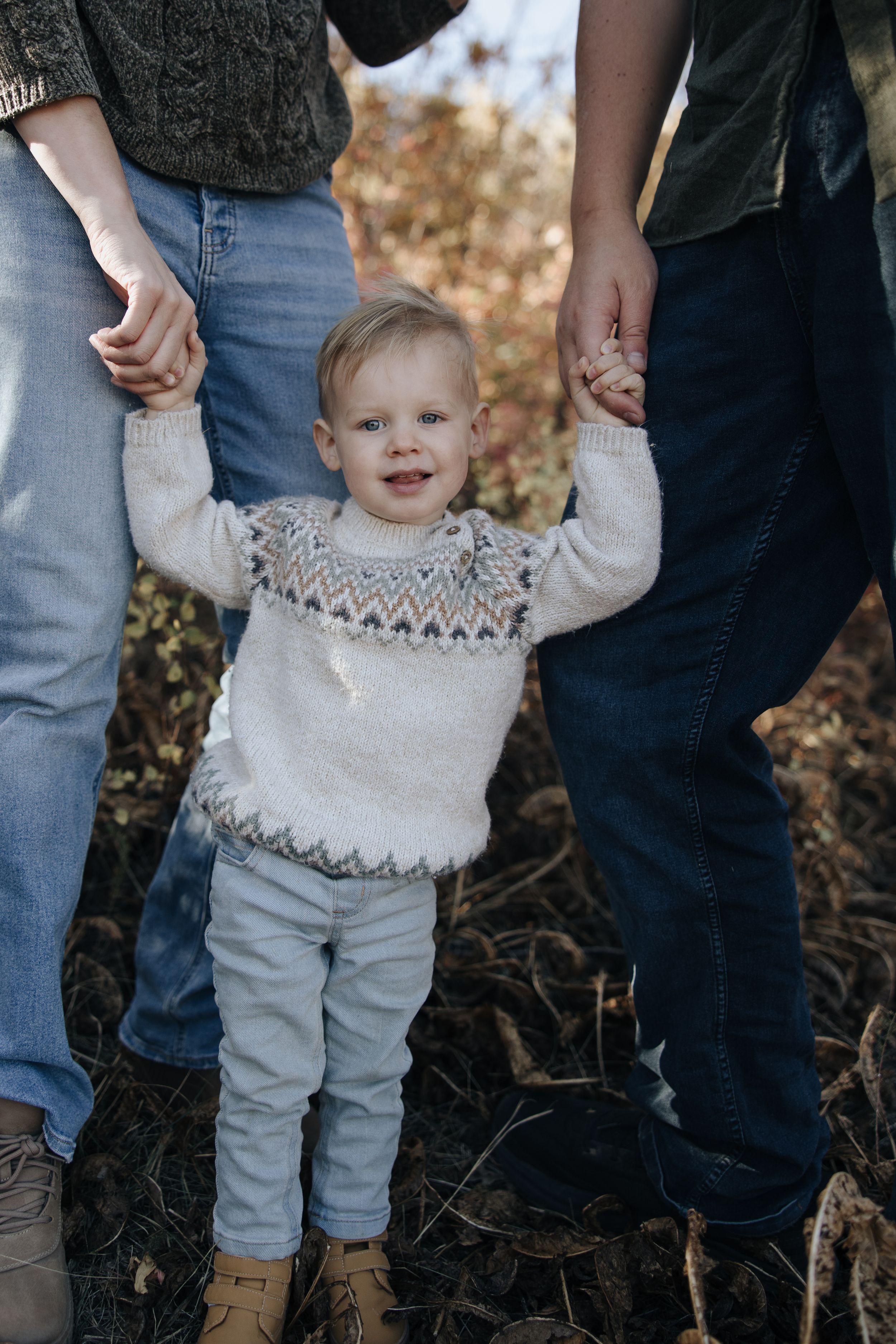A small child in a cozy knit sweater holds hands with parents while standing outdoors on an autumn day.