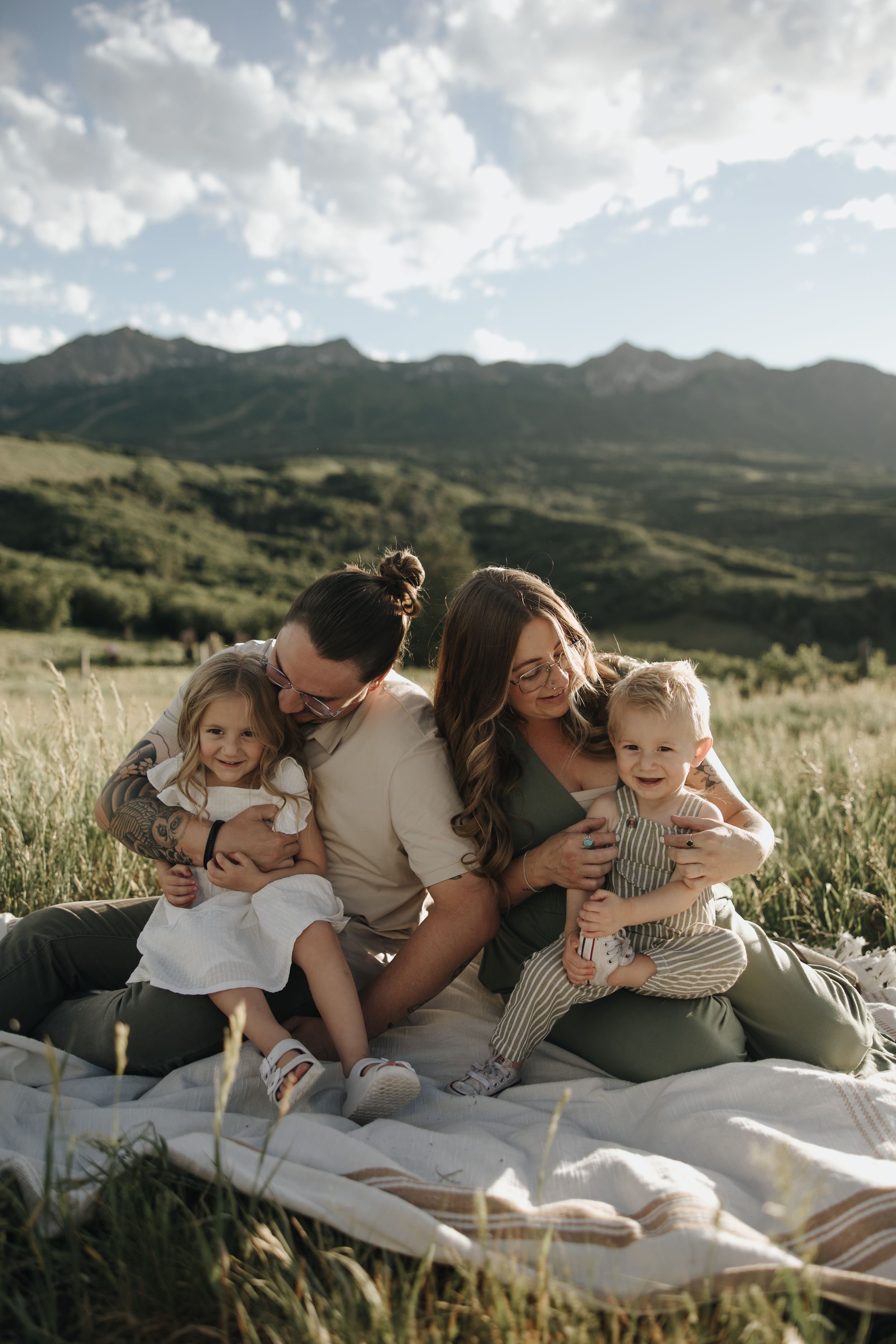 Family enjoys a picturesque picnic on a blanket in a grassy meadow with mountain landscape and cloudy sky backdrop.
