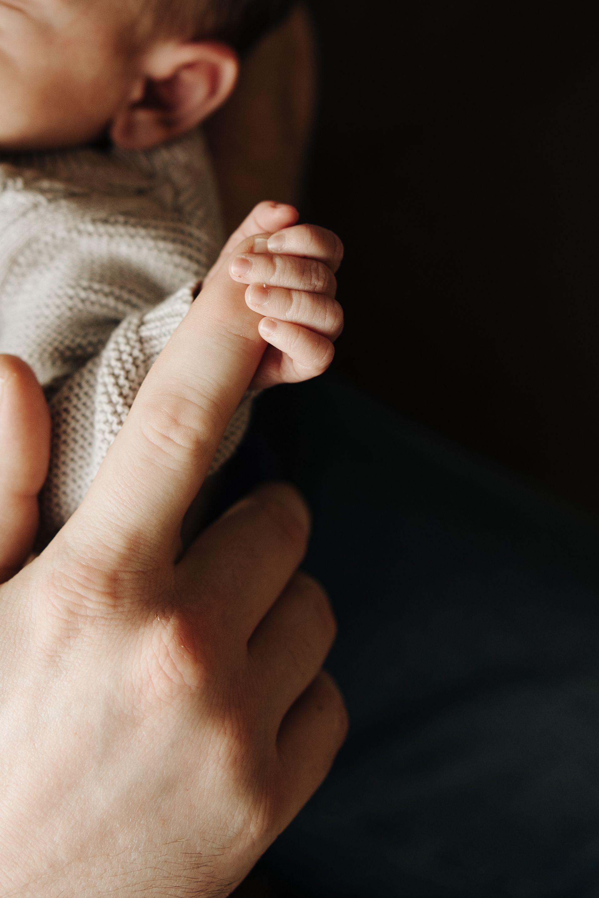 Close-up shot of tiny baby fingers grasping an adult's finger while wearing a knit sweater on dark background.