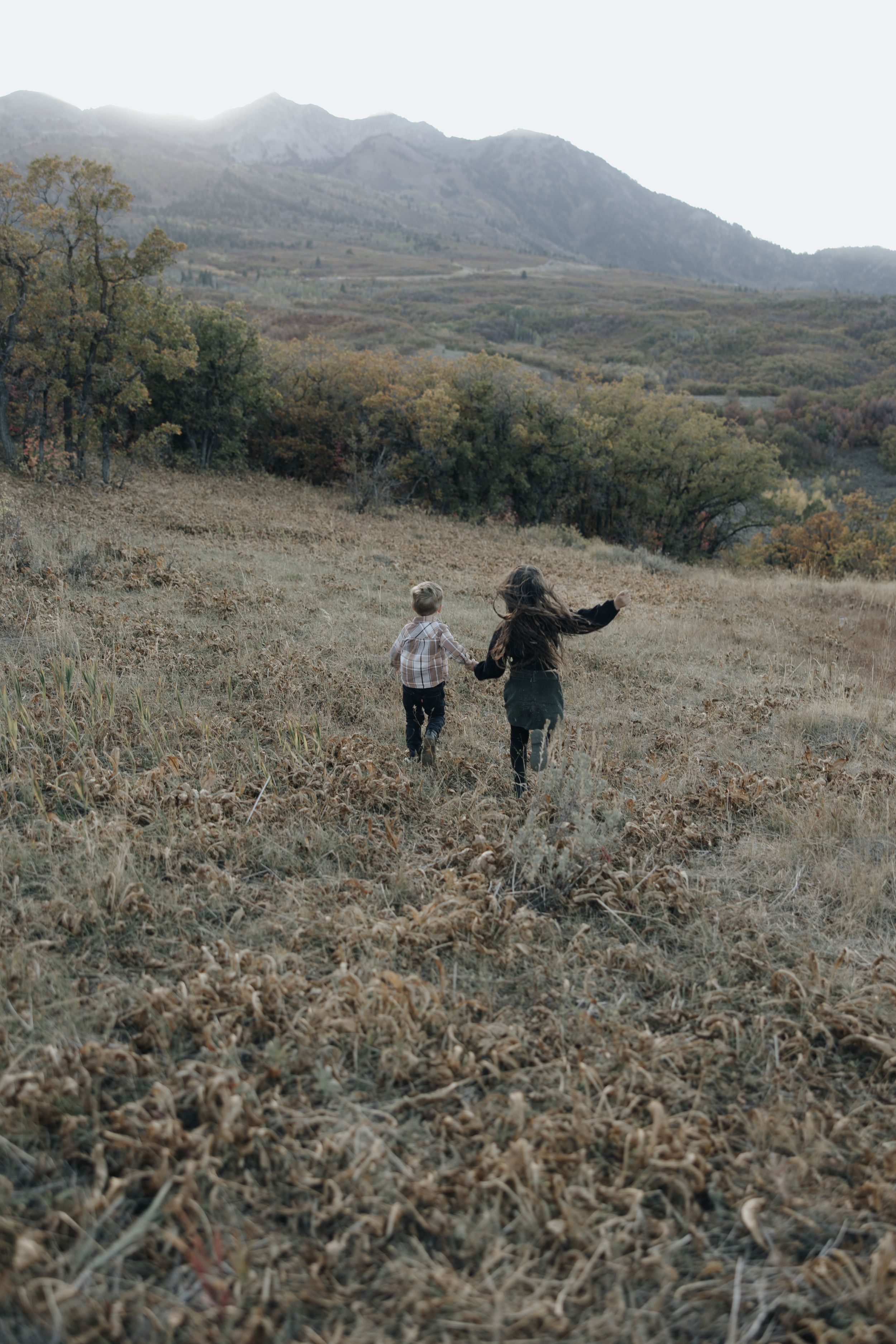 Two people walking together on a rugged mountainside trail through dry grass with mountains in the distance.