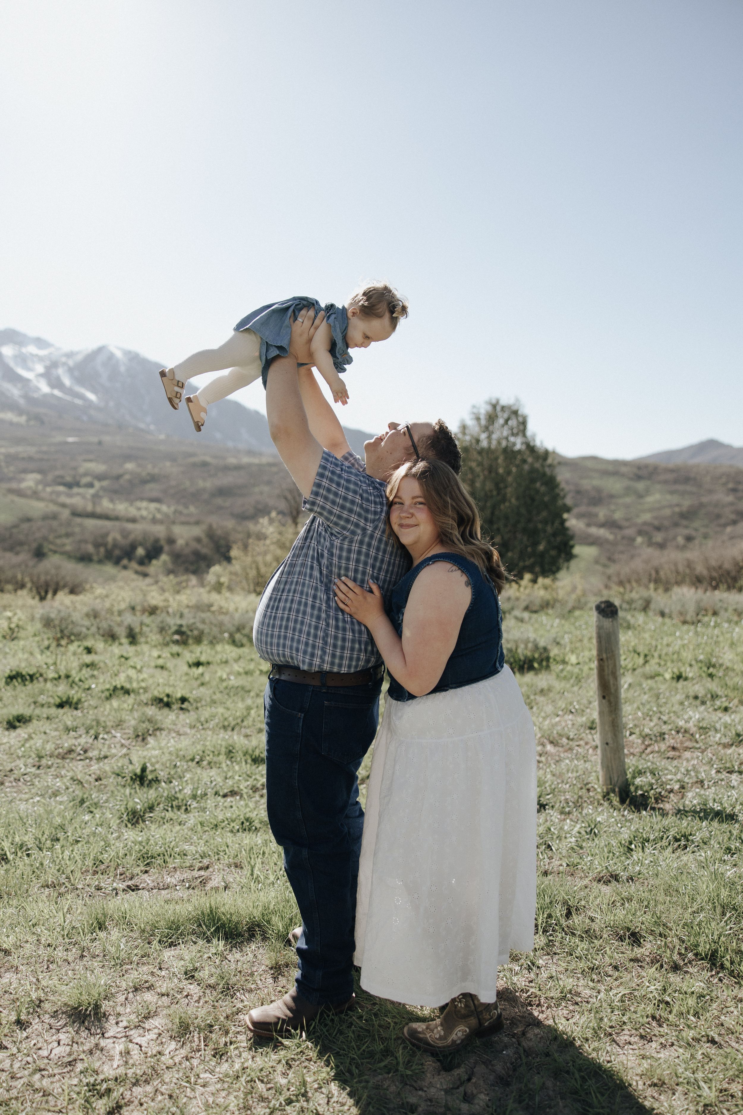 A couple stands in a scenic mountain field playing with a small child against a backdrop of rolling hills and clear skies.