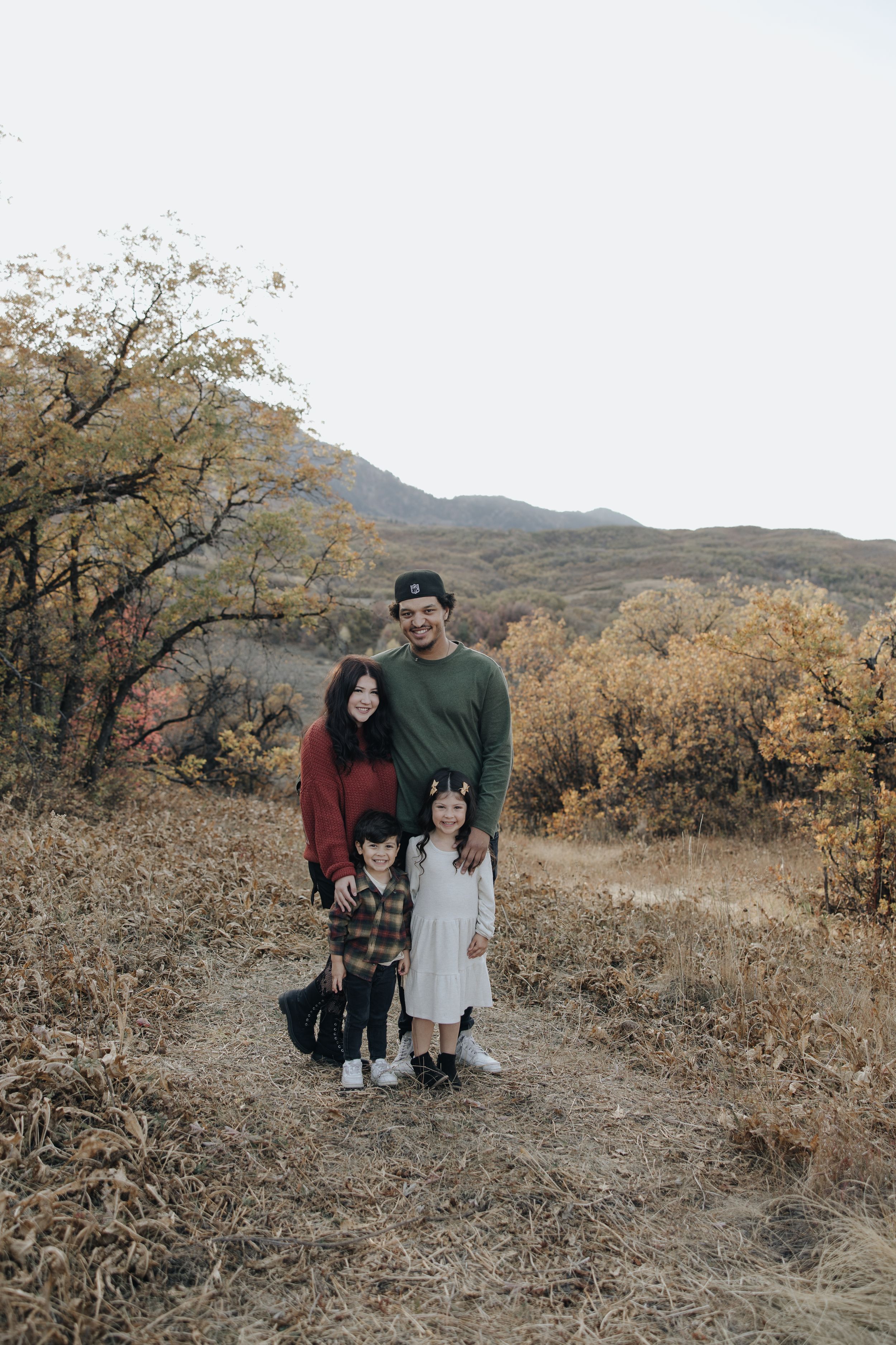 Family standing together on autumn mountain trail surrounded by golden fall foliage and hills in background.
