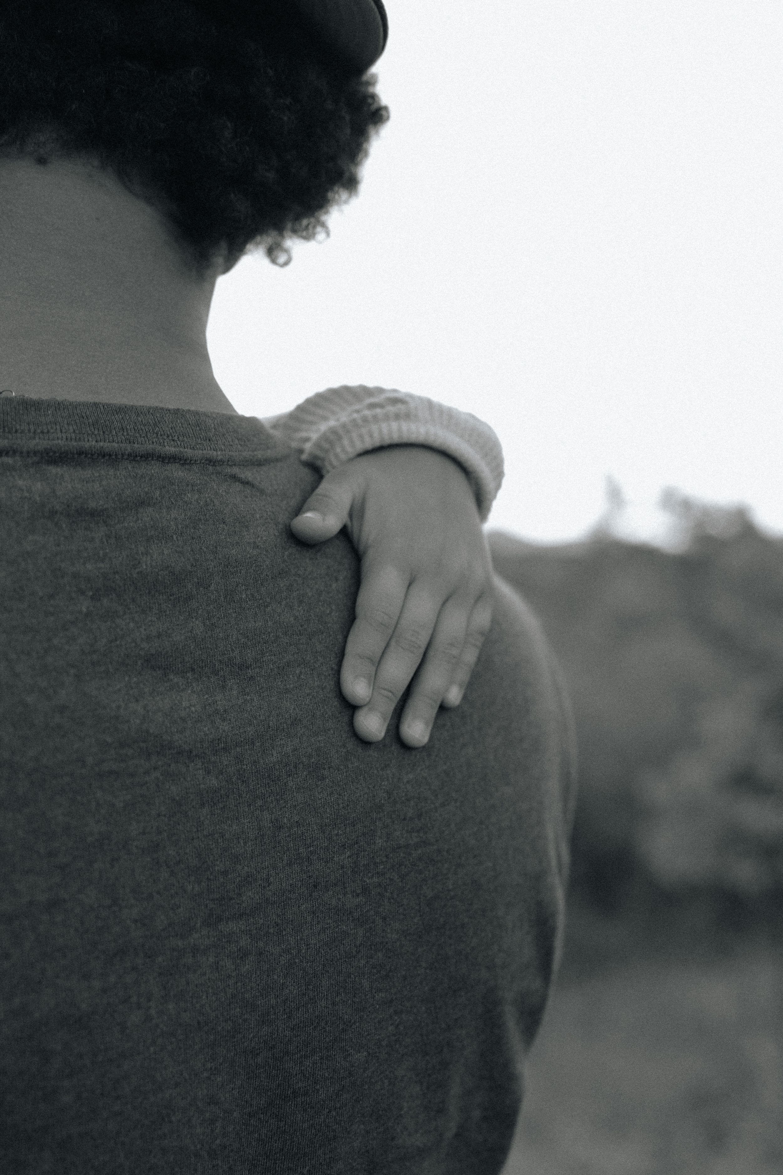 A comforting hand rests on someone's shoulder wearing a gray shirt, creating an intimate black and white composition.